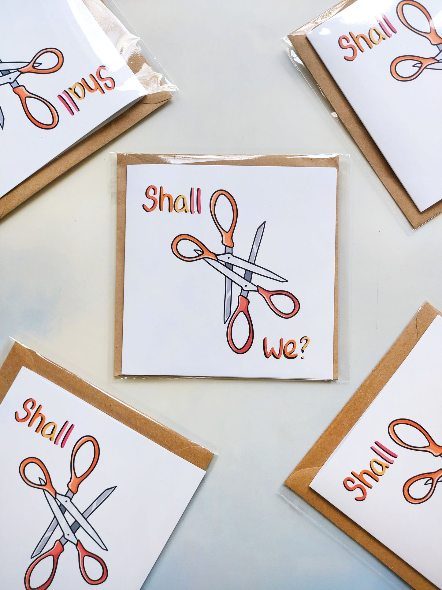 Shall We Scissor? • Funny Lesbian Greeting Card - EmilsDoodles
On a sky background are five packaged greeting cards. On the cards are two interlocking scissors, with the phrase: shall we?, on it. All in the colours of the lesbian pride flag.