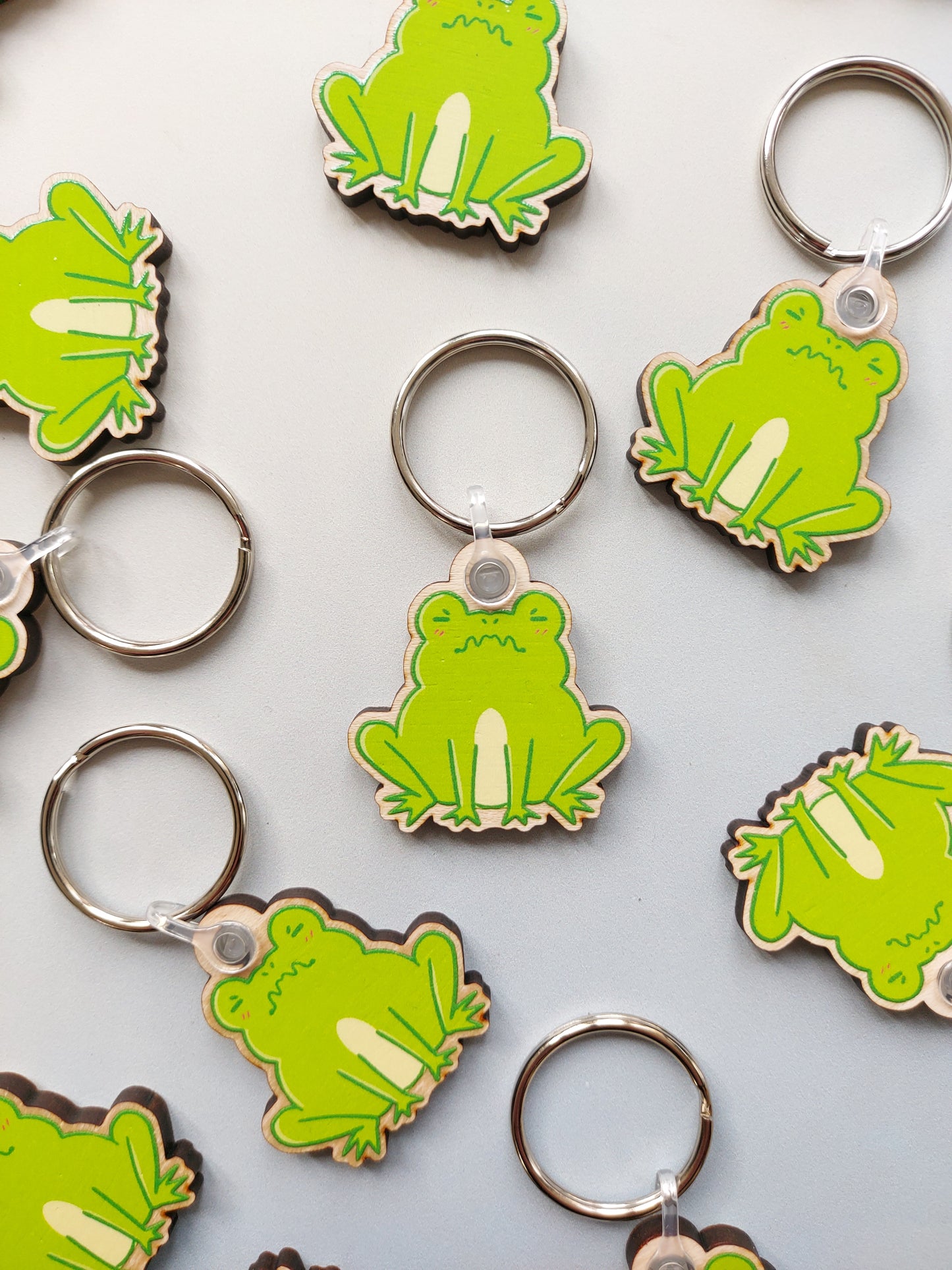 On a light grey background, several of the same keyring design are scattered around. The keyring is made from wood and has a design of a grumpy frog. The outline of the design is a dark green. The frog is colour a light green with a yellow patch between the two front legs, underneath both eyes are two pink dashes for blush.