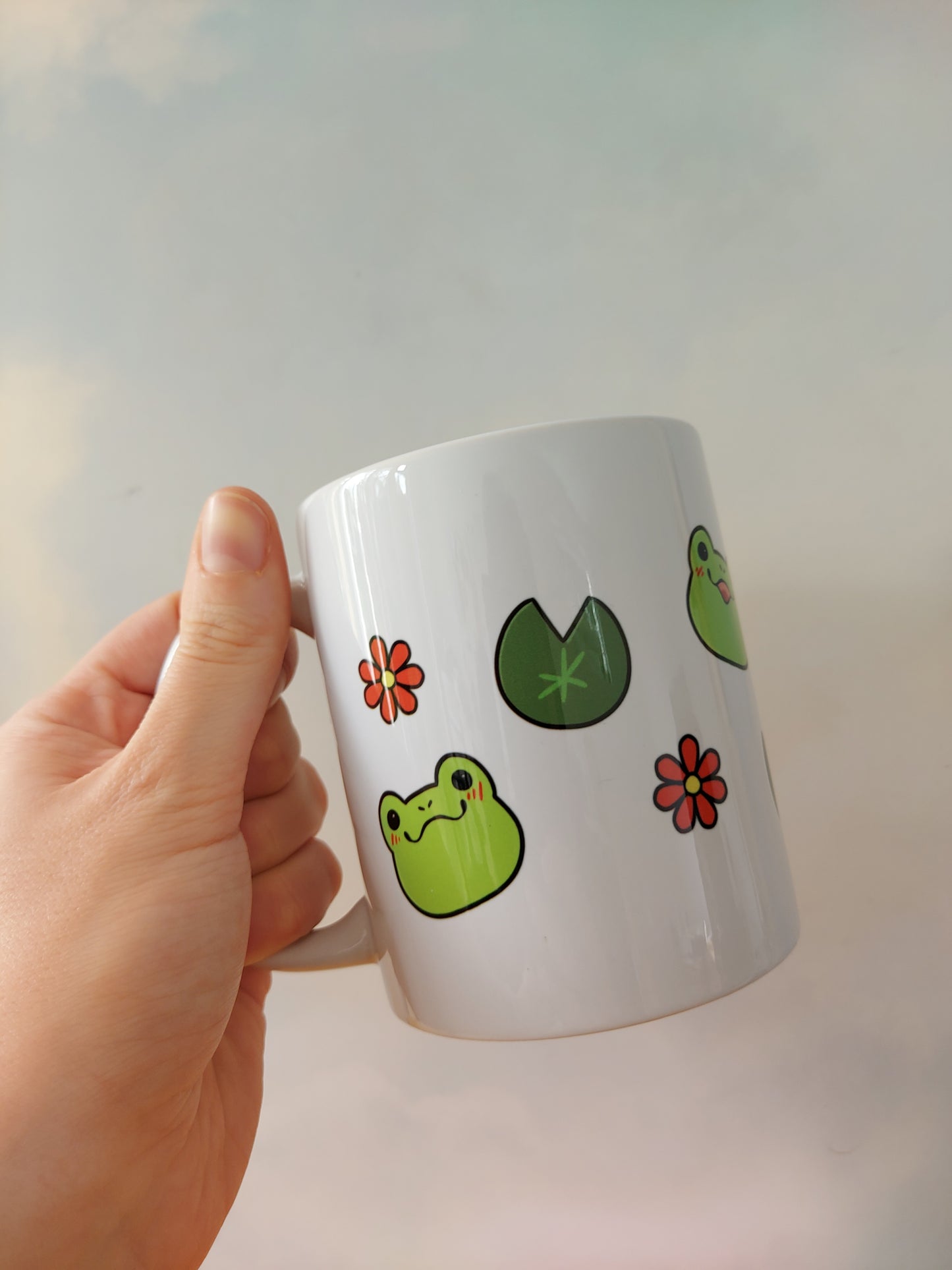 Emotions of a Frog 11oz Mug - EmilsDoodles
A hand holding a mug. On the mug are frogs, flowers, and lily pads. Each frog face has a different emotion. They're also drawn in a kawaii art style.