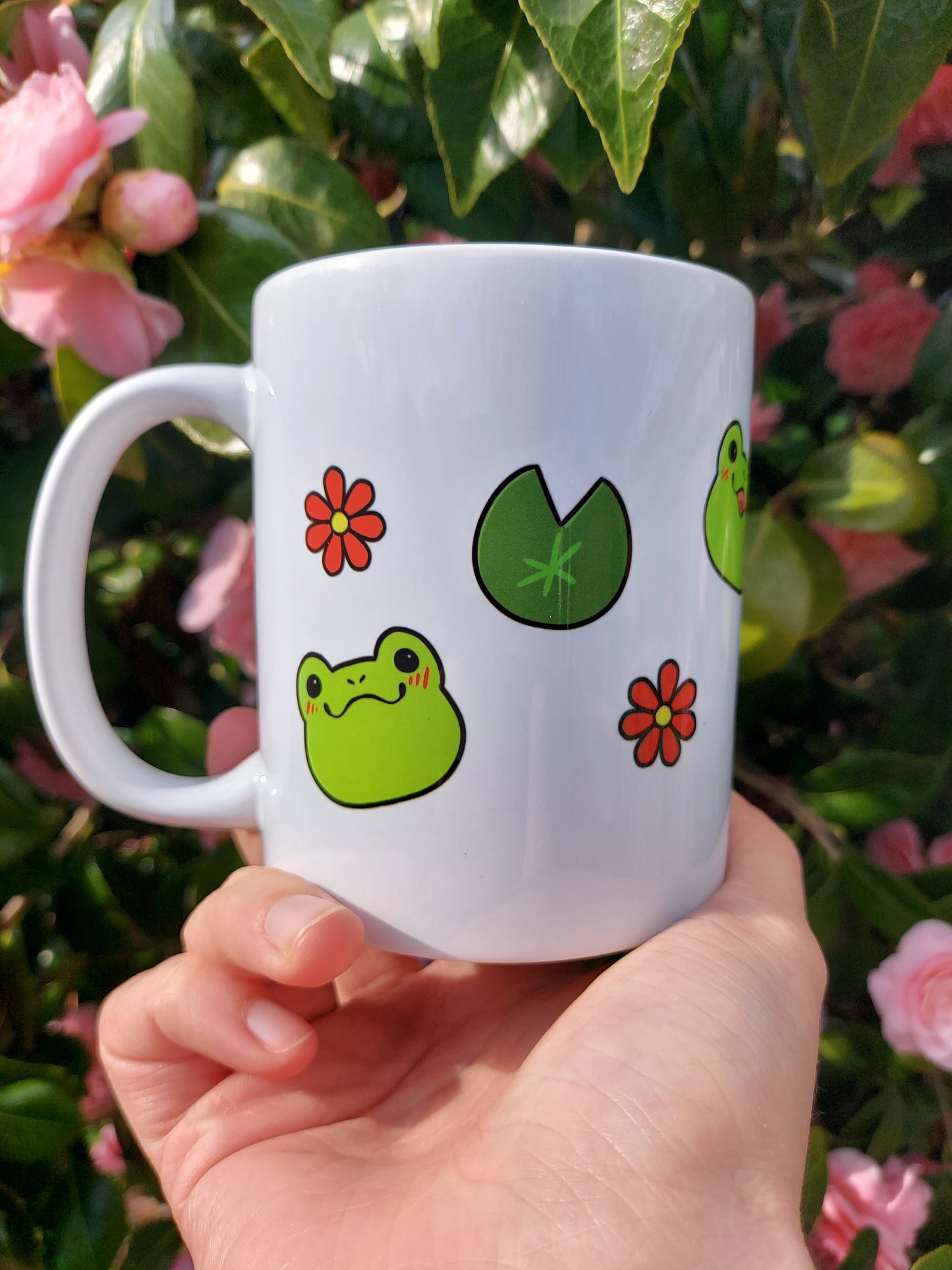 Emotions of a Frog 11oz Mug - EmilsDoodles
In front of flowers, a hand holds a mug with kawaii frogs, flowers, and lily pads.