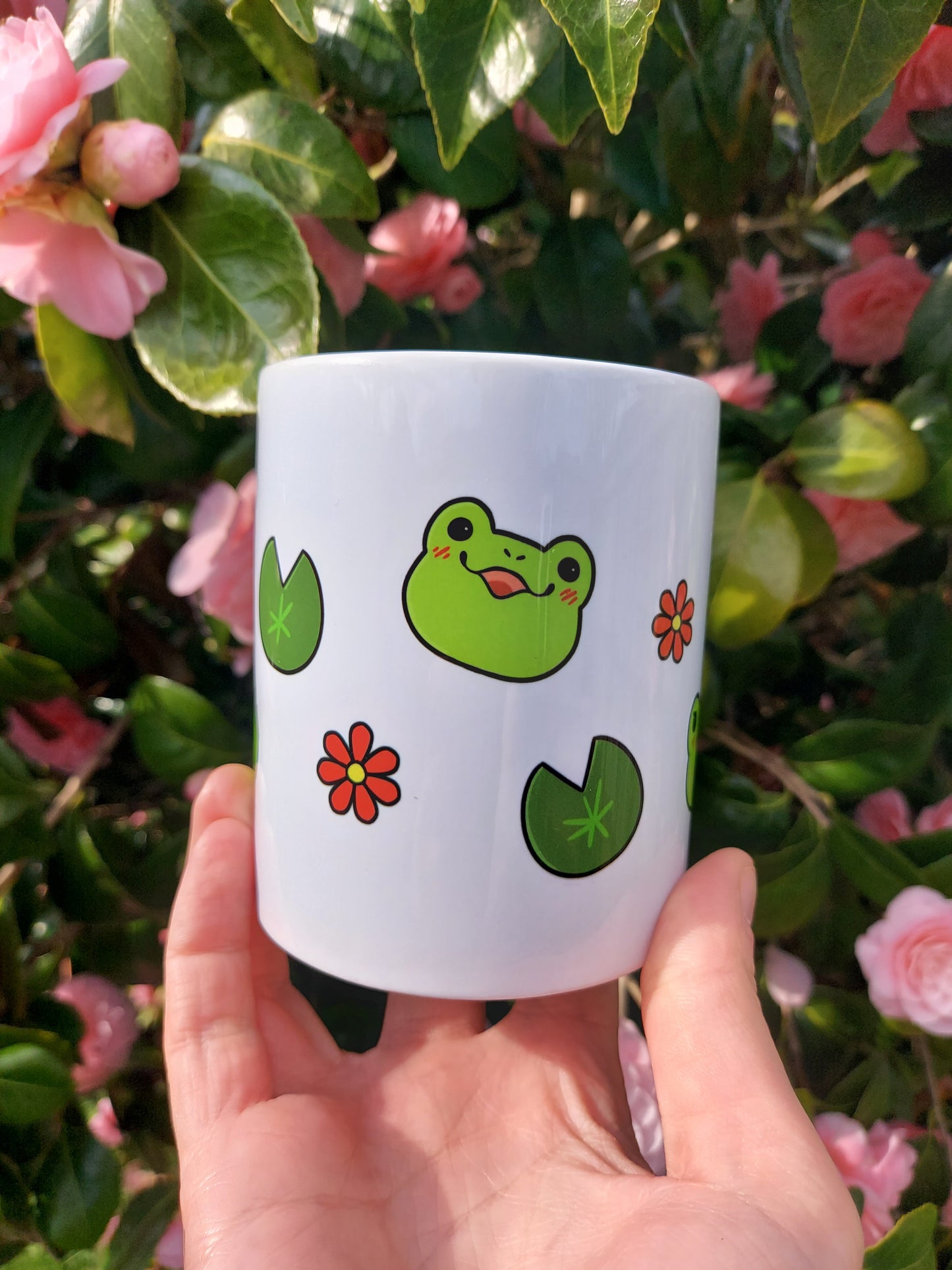 Emotions of a Frog 11oz Mug - EmilsDoodles
In front of flowers a hand holds a mug. There is a kawaii frog with a smiling expression. Around it are lily pads and flowers.