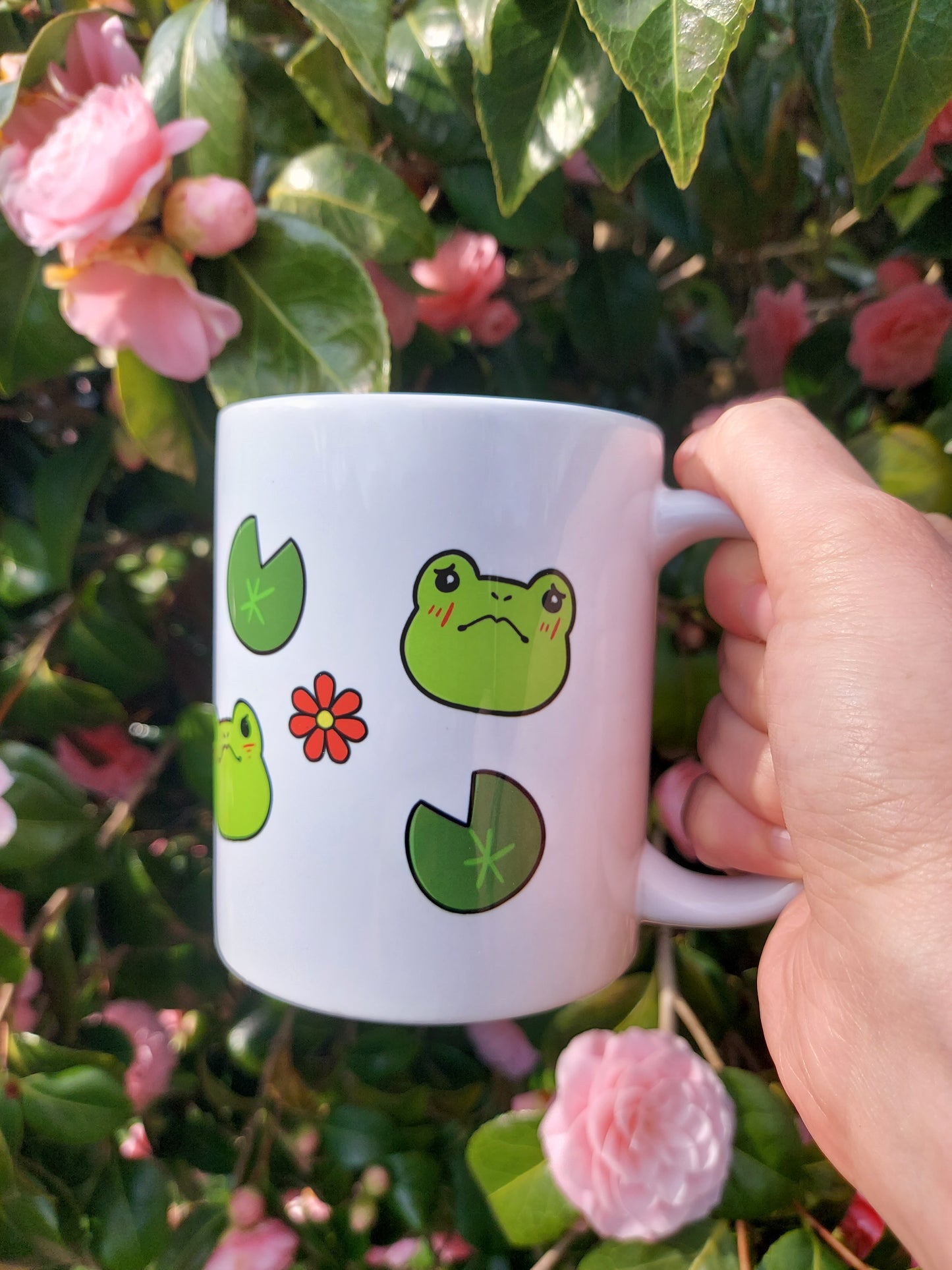 Emotions of a Frog 11oz Mug - EmilsDoodles
In front of flowers, a hand holds a mug with cute frogs, lily pads, and flowers. One of the frogs has a sad look on its face.