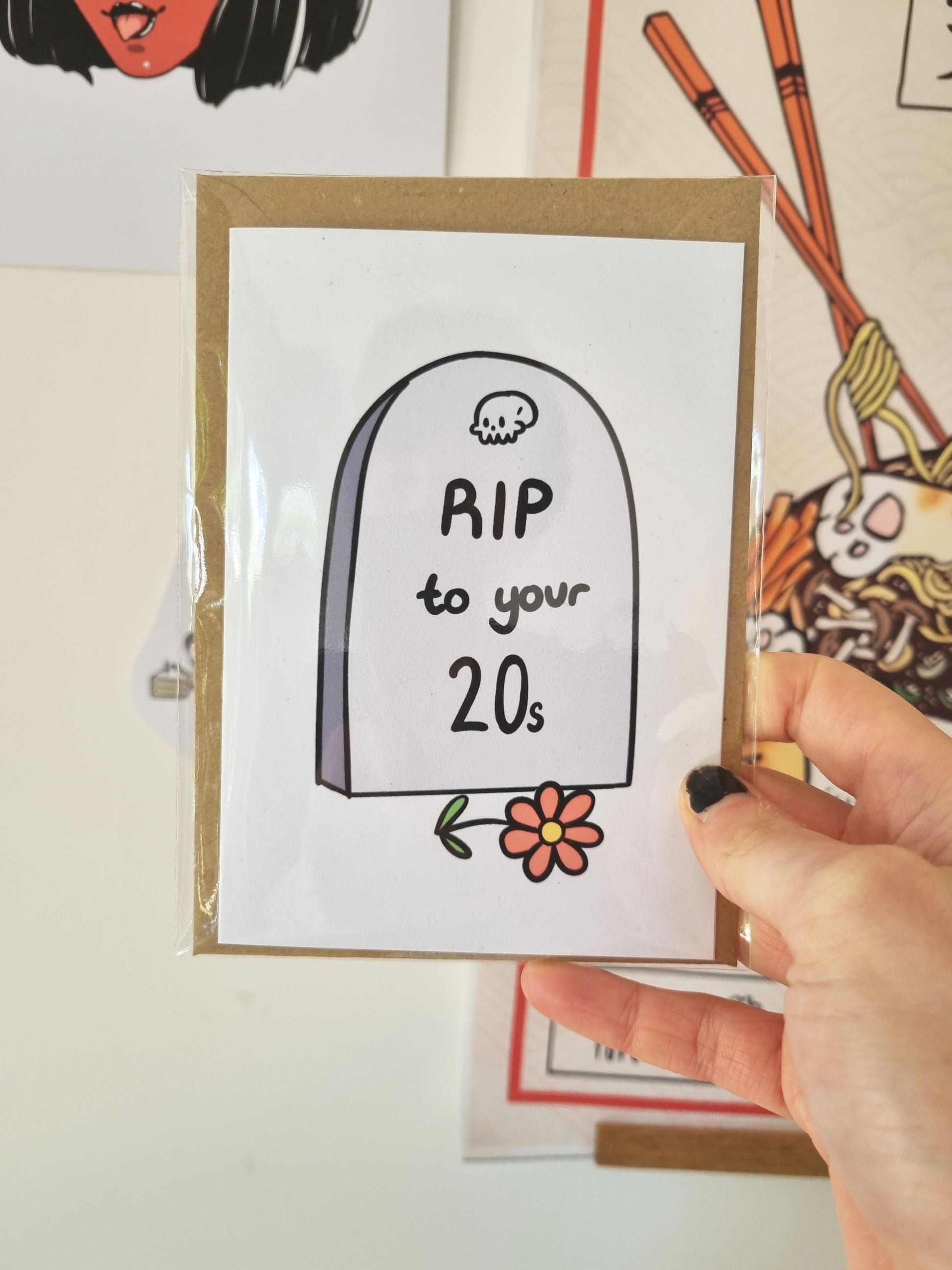 A hand holds a packaged greeting card. The card is A6 in size and behind is a kraft envelope. The greeting card has a tombstone, at the top is a cute skull drawing. Underneath is written: RIP to your 20s. In front of the headstone is a pink flower.
