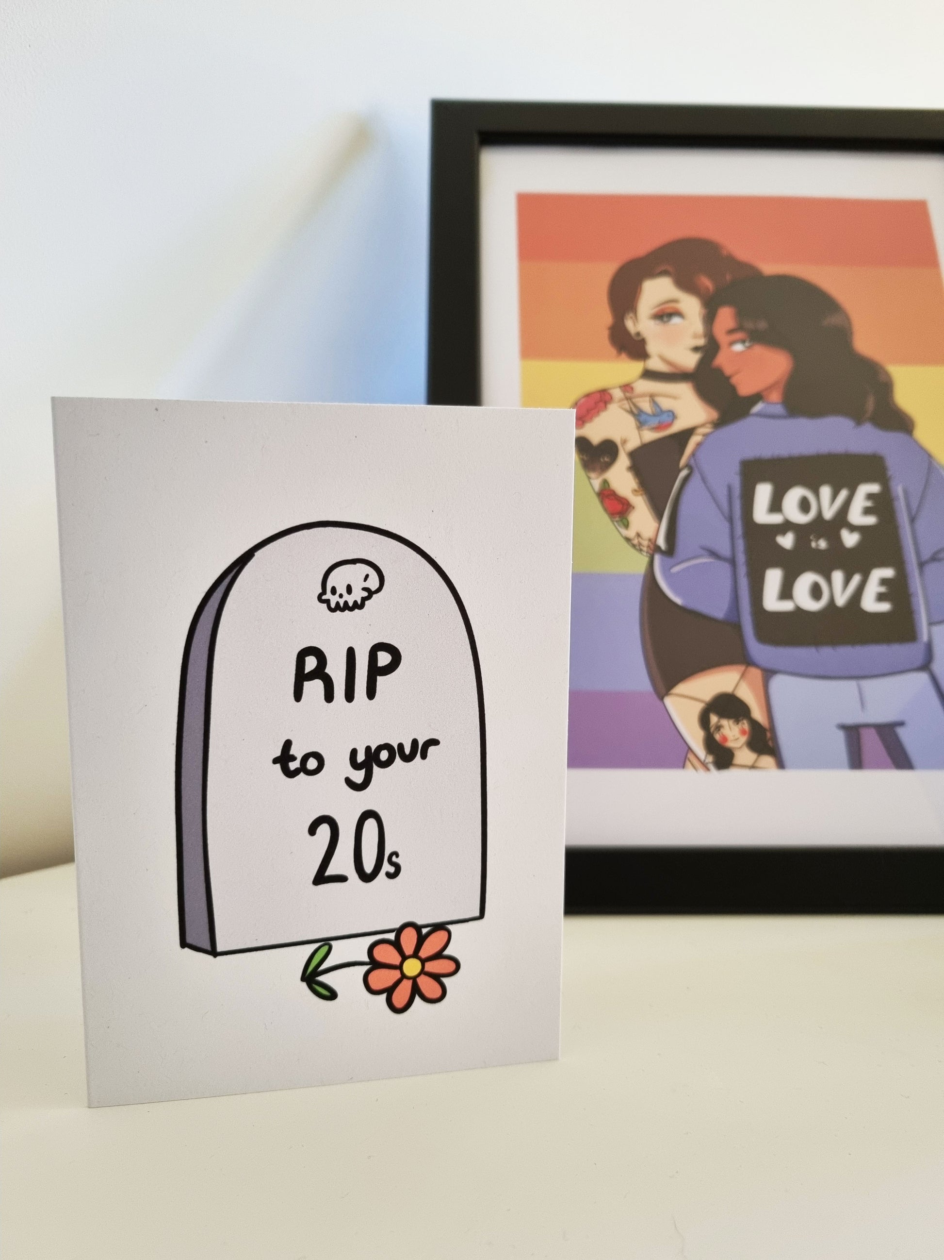 The RIP to your 20s greeting card is standing in front on top of a white table, with a print in the background.