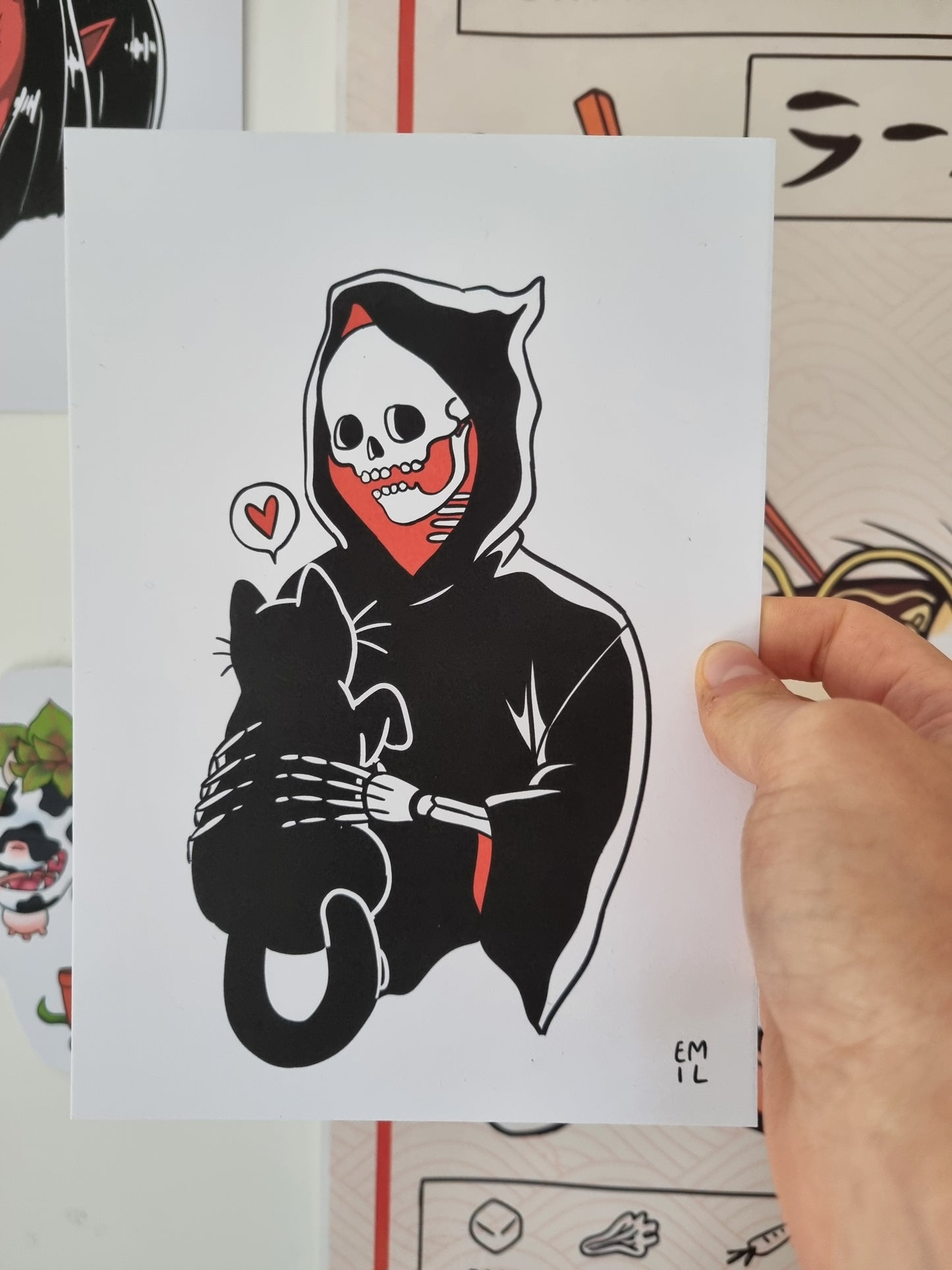 A hand is holding an A5 art print. The art print is of a cute grim reaper. The inside of the grim reapers cloak is red. In his hands is a black cat. From the black cat is a speech bubble with a love heart inside.