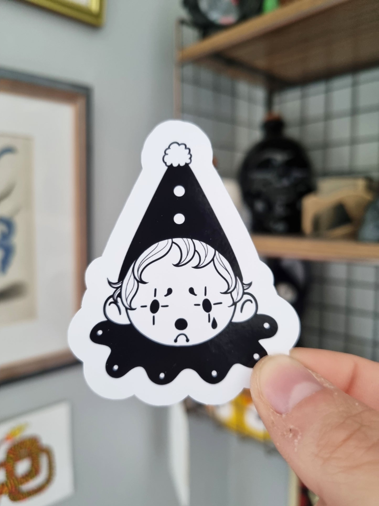 The Sad Clown | Glossy Clown Sticker - EmilsDoodles
A hand holds a glossy sticker with a design of a sad clown. It's black and white. The hat and ruffle is black.