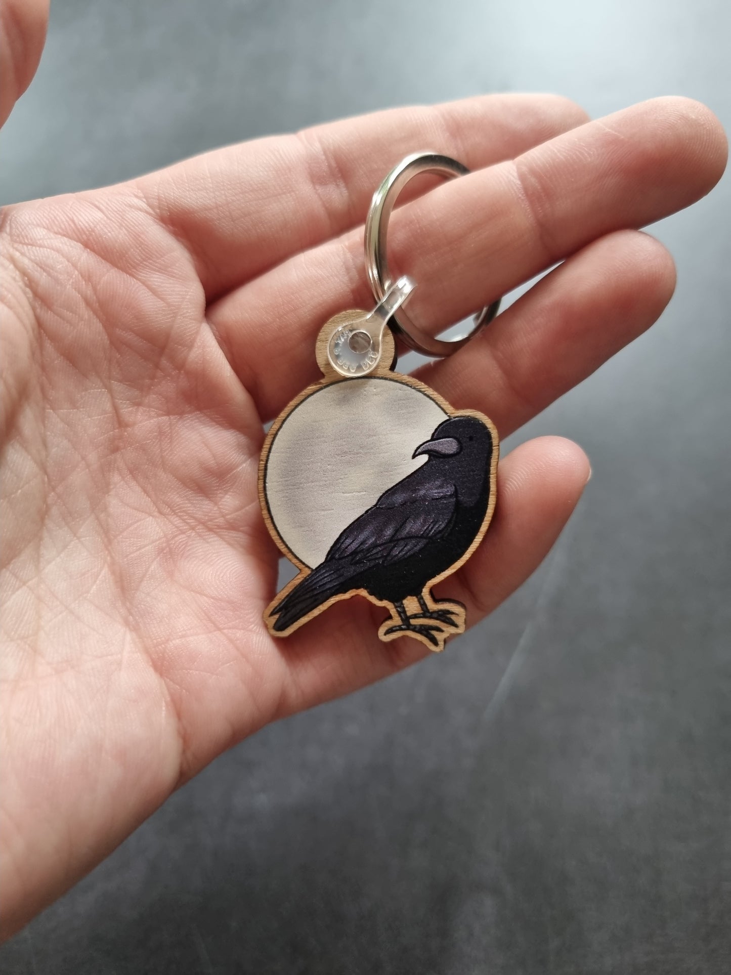 A hand holding a wooden keyring. The keyring is a raven in front of the moon.