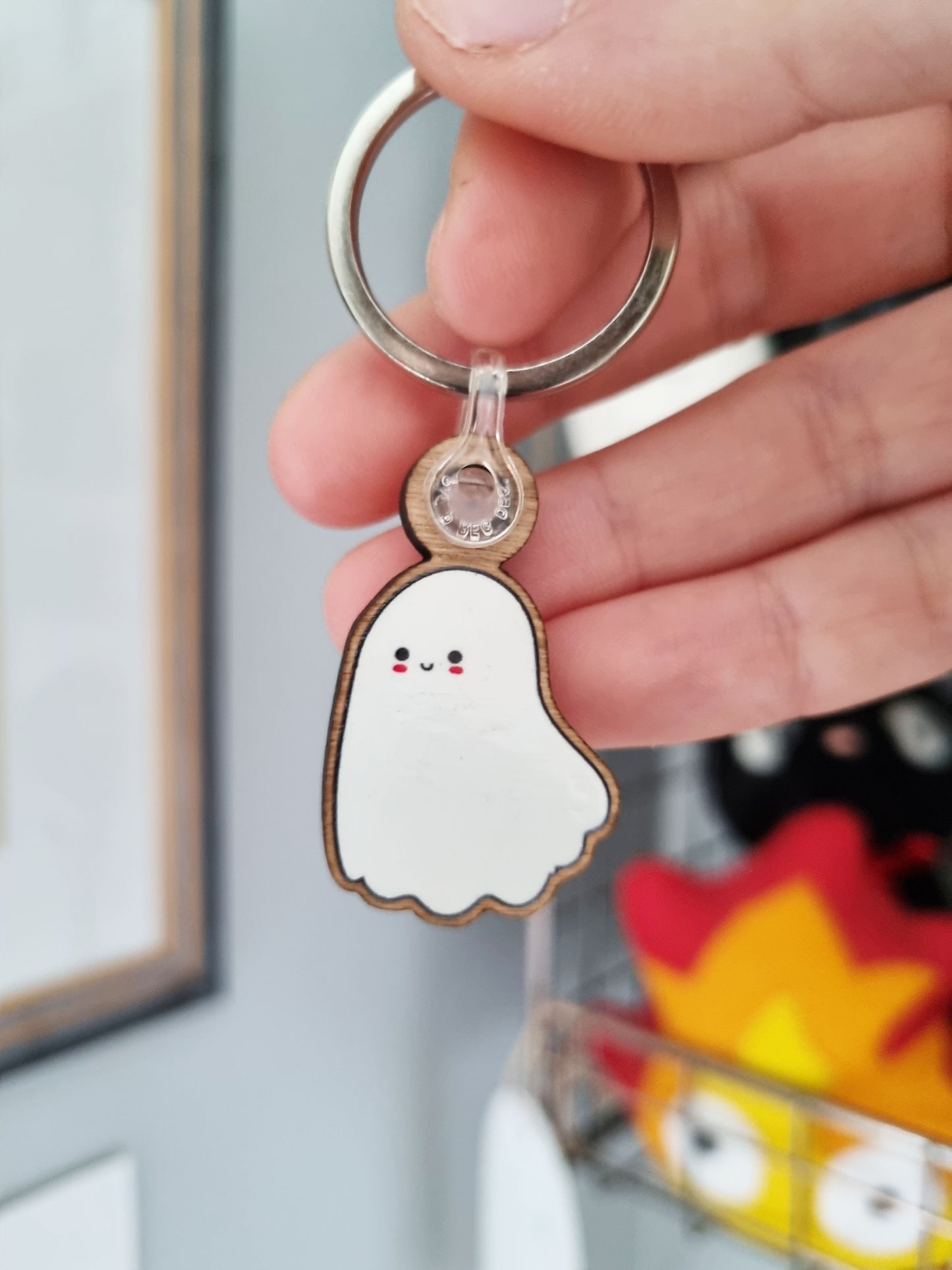 A hand holds a cute ghost keyring made from wood. The ghost is smiling and has pink circles underneath the eyes to look like blush.