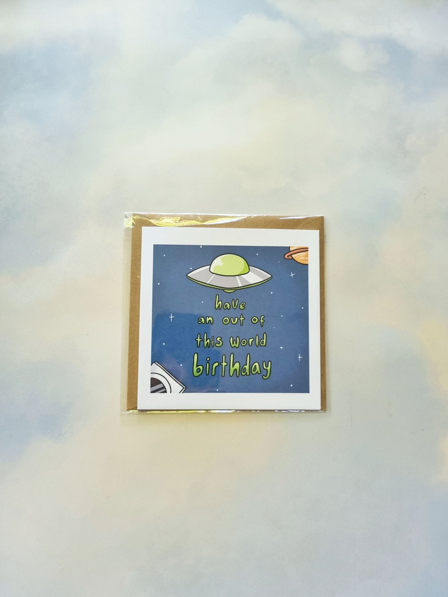 On a sky background, is a packaged greeting card. On the greeting card is a space scene with a UFO above the phrase: Have an out of this world birthday.
In the bottom left corner is an astronaut, and in the top right corner is a ringed planet.