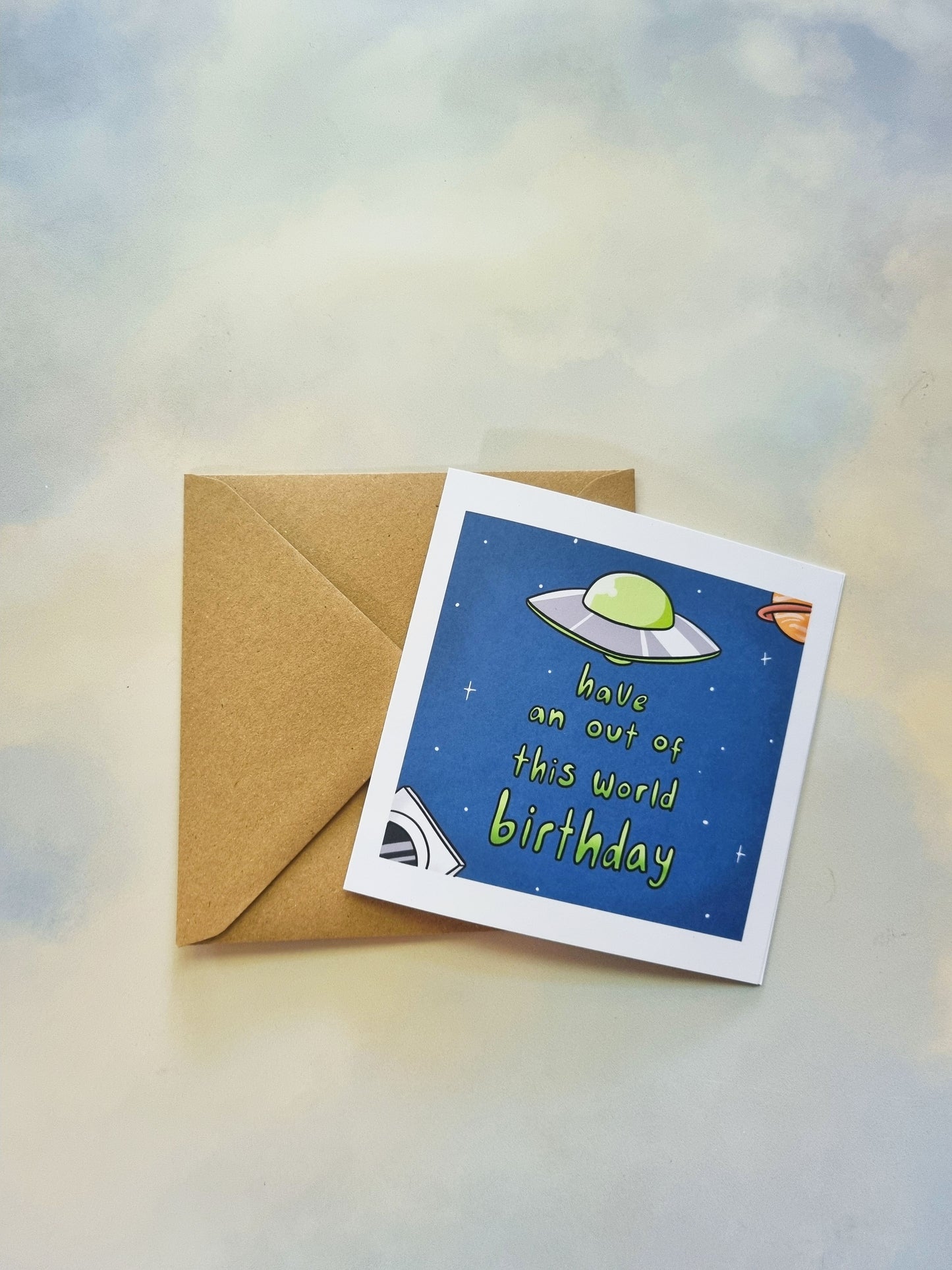 On a sky background is a kraft envelope and a greeting card. On the greeting card is a space scene with a UFO above the phrase: Have an out of this world birthday.
In the bottom left corner is an astronaut, and in the top right corner is a ringed planet.