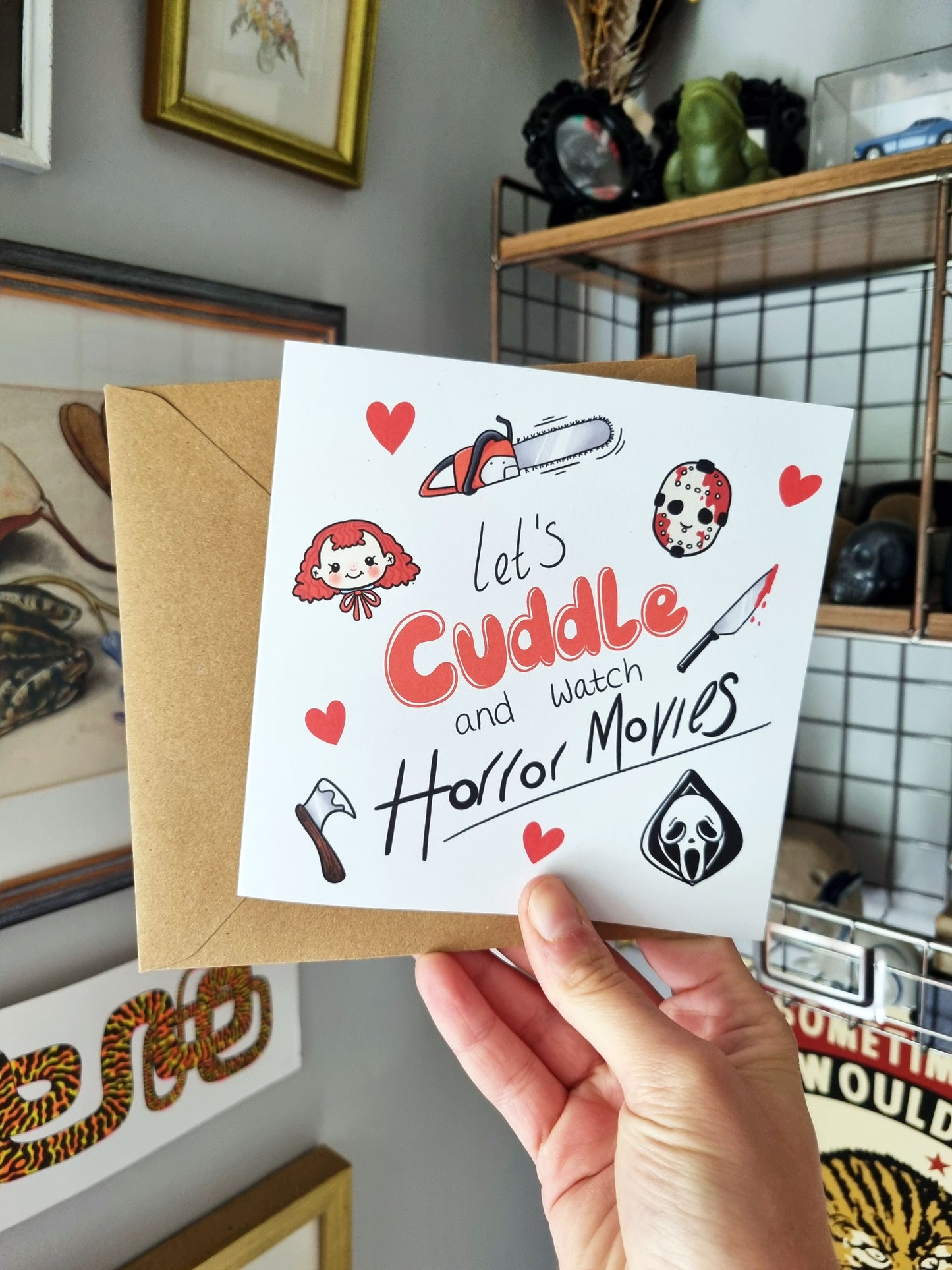 A hand holding a square greeting card and kraft envelope. The greeting cards reads: Let's cuddle and watch horror movies. Around this are horror icon inspired drawings. A raggedy Ann doll head, a bloody hocky mask, scream mask, chainsaw, bloody knife and a hatchet, as well as love hearts.