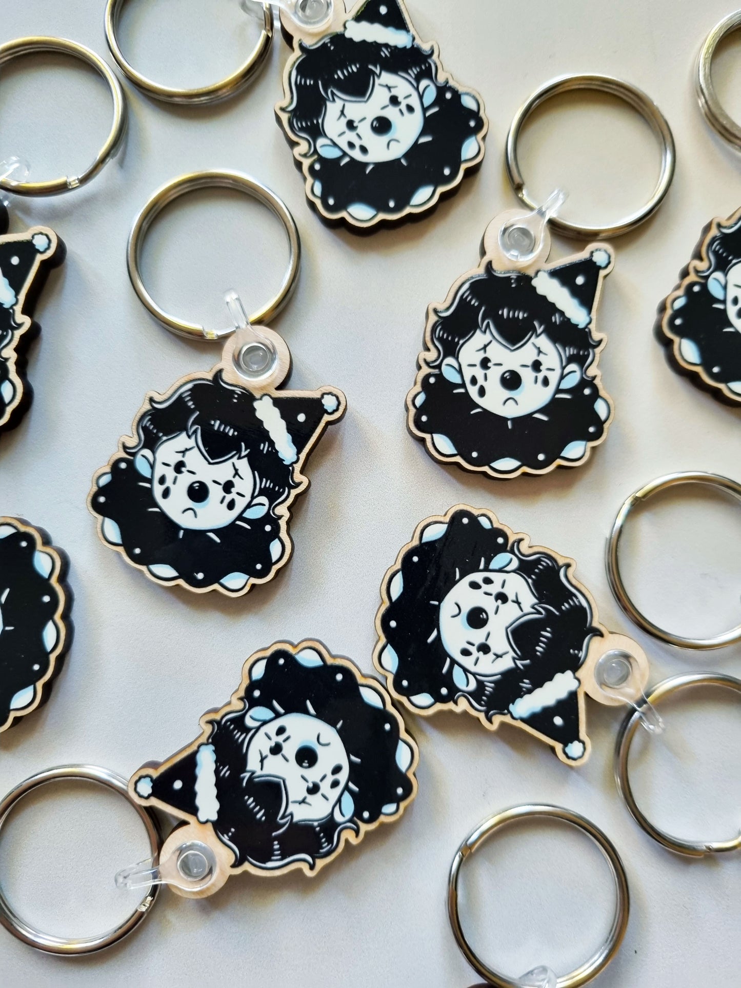 On a white background, the same design of clown keyrings are randomly scattered around. The clown design is black and white and has a sad face. Underneath the eyes are black tears. The clown wears a black ruffle with a white ruffle under neath that. And at an angle on it's head wears and cone hat with white ruffles on the top and bottom. The keyring is made from wood and has a circler attachment at the top,