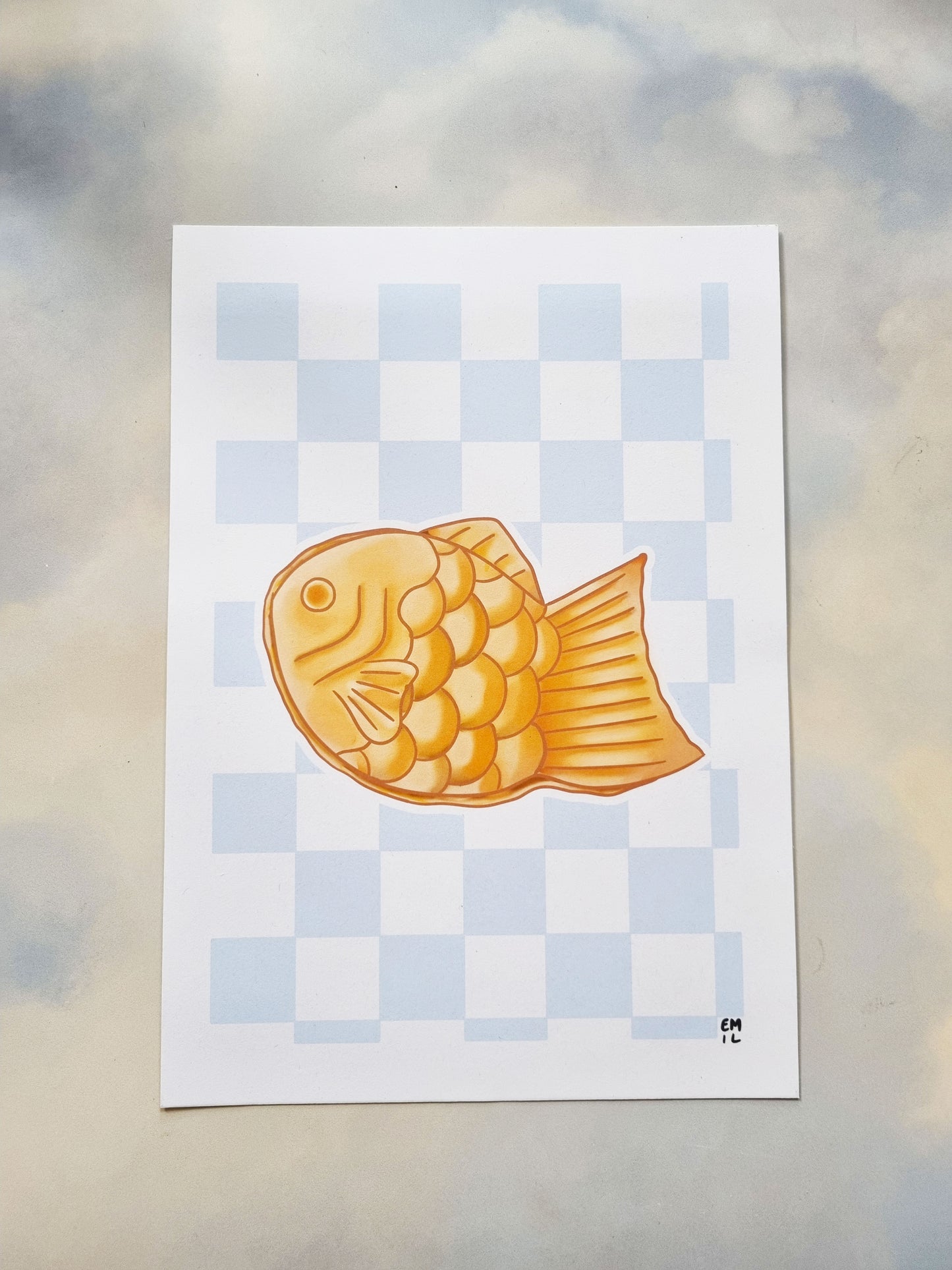 On a sky background lays an A4 art print. On the art print is a light blue checked pattern. On top of this is a Japanese desert called Taiyaki, which is a waffle filled with custard or red bean paste in the shape of a fish.