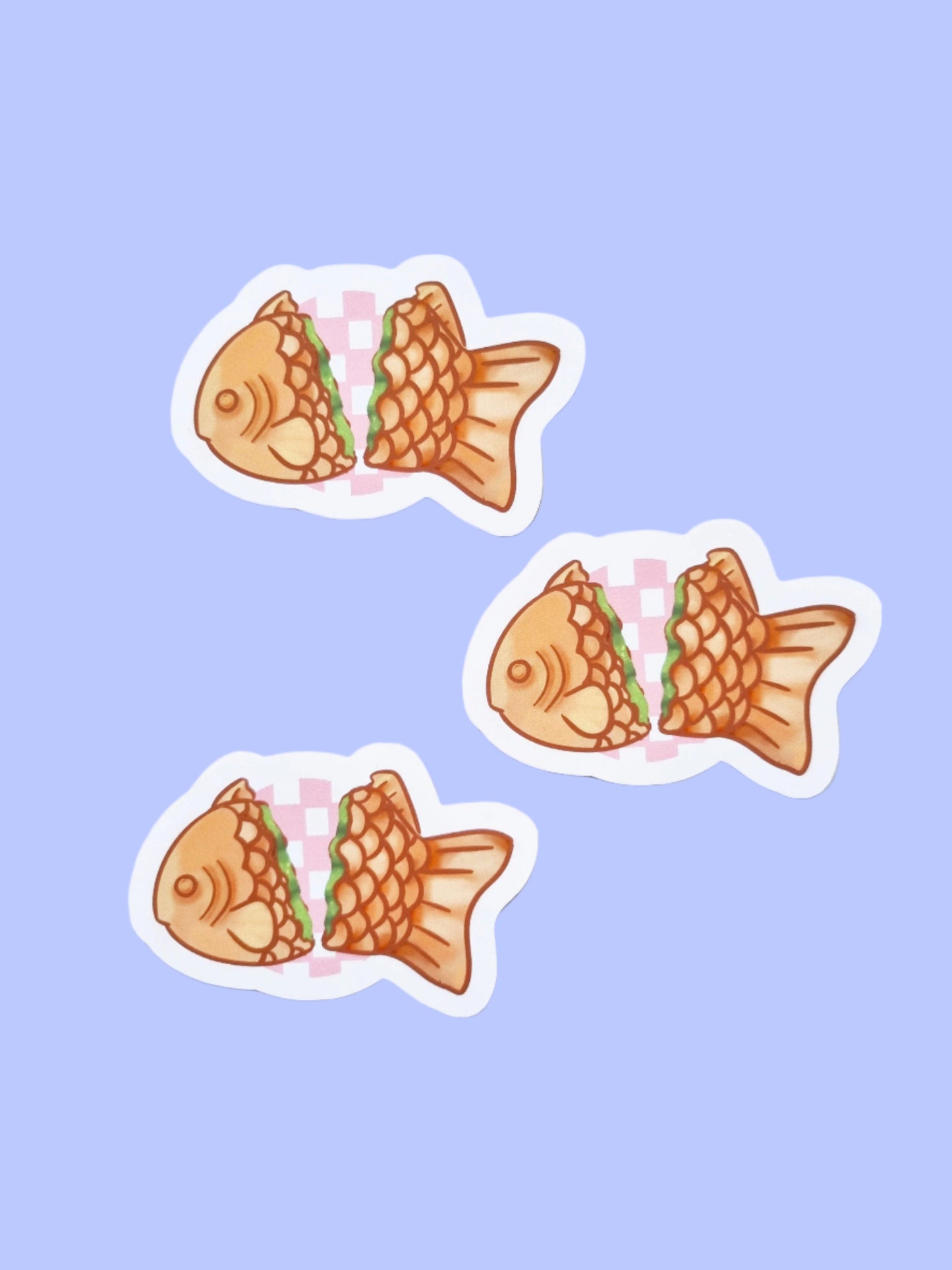 On a periwinkle background are three stickers. The stickers are of a Japanese desert called taiyaki. The fish shaped sticker is split in two to show a matcha filling. Between the two sides are a circler, pink checked background.