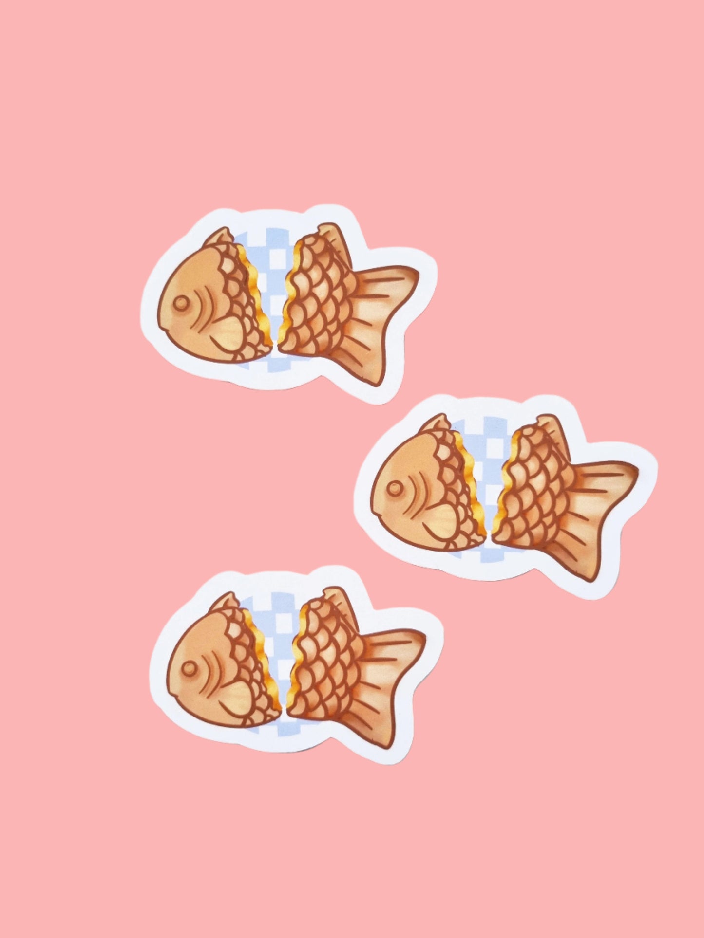 On a pink background are three stickers. The stickers are of a Japanese desert called taiyaki. The fish shaped sticker is split in two to show a custard filling. Between the two sides are a circler, light purple checked background.