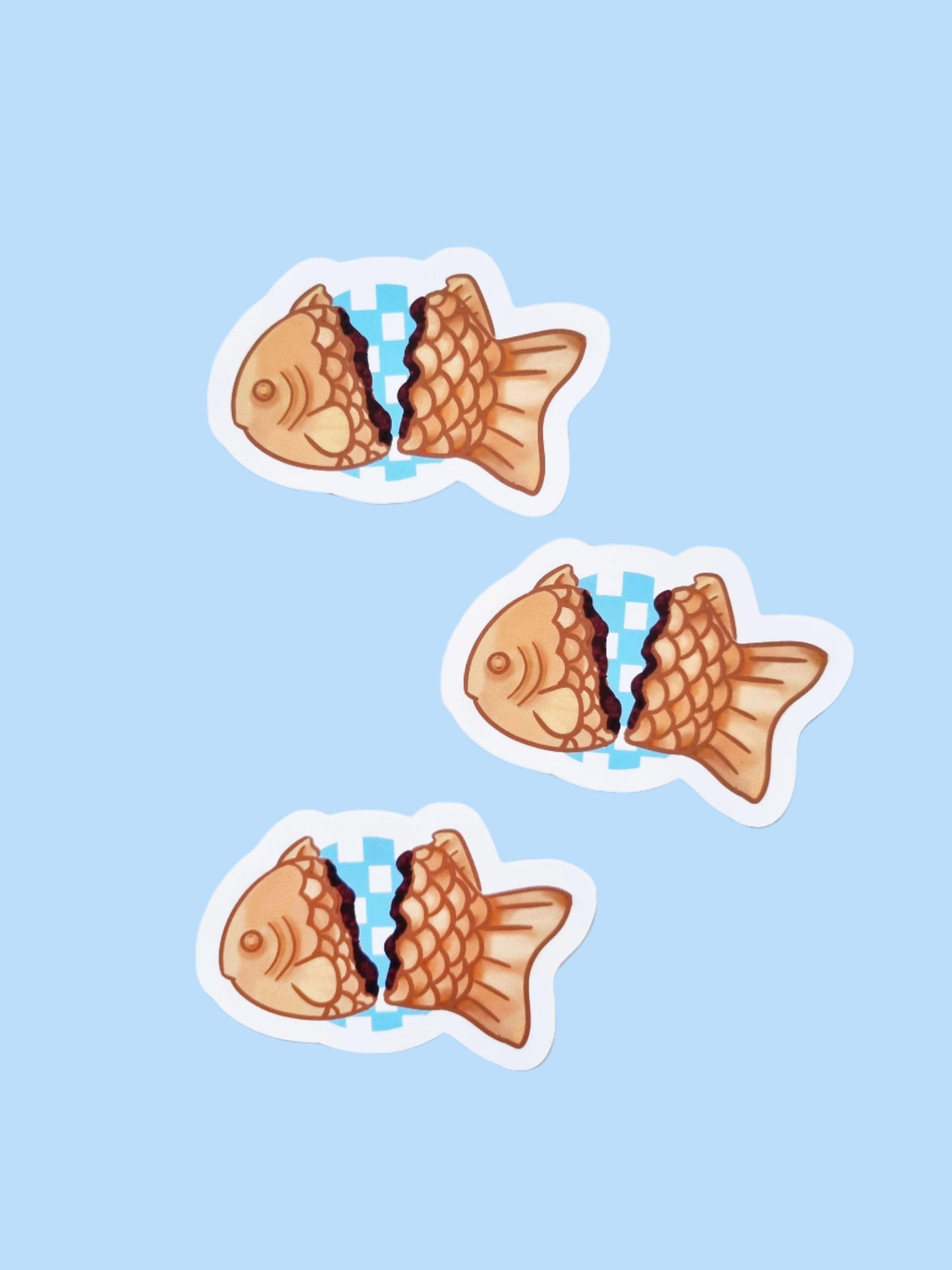 On a light blue background are three stickers. The stickers are of a Japanese desert called taiyaki. The fish shaped sticker is split in two to show a red bean filling. Between the two sides is a blue checked circler background.