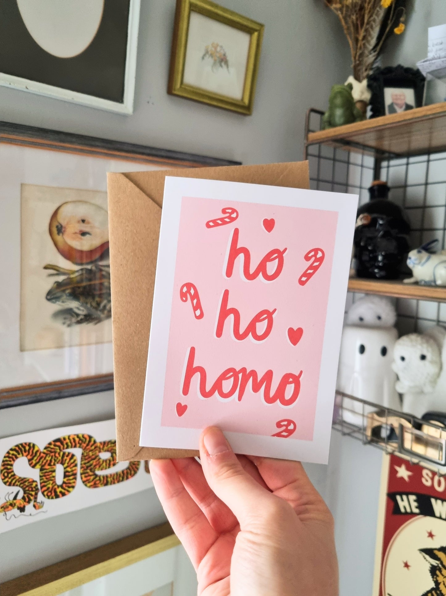 A hand holding an A6 card, with a pastel pink background and white border. Inside the pastel pink background is the phrase: Ho, Ho, Homo, written in a red cursive font. Around the phrase are candy canes and love hearts.