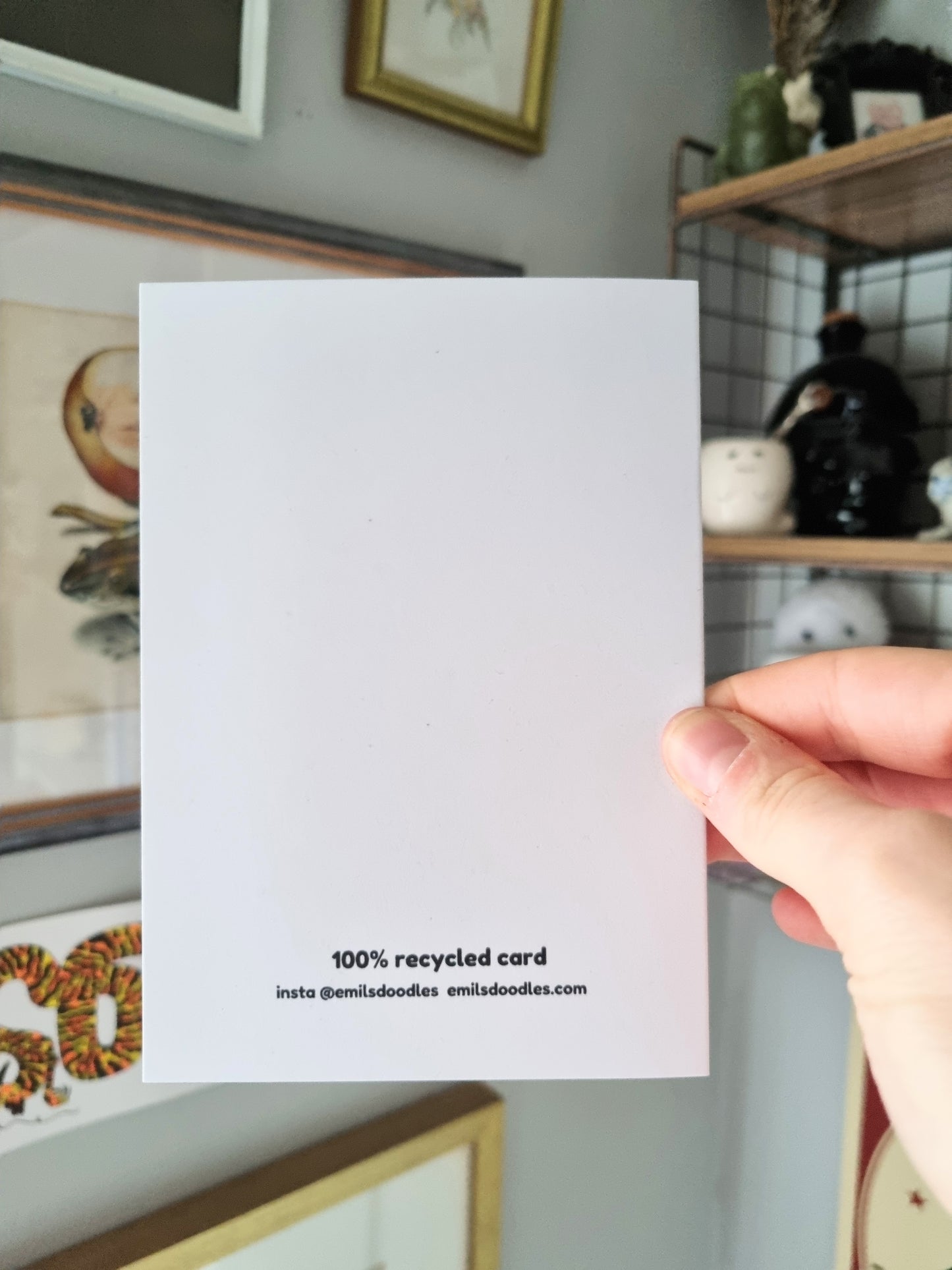 A hand holds a greeting card showing the back of it. It has the shops branding on it.