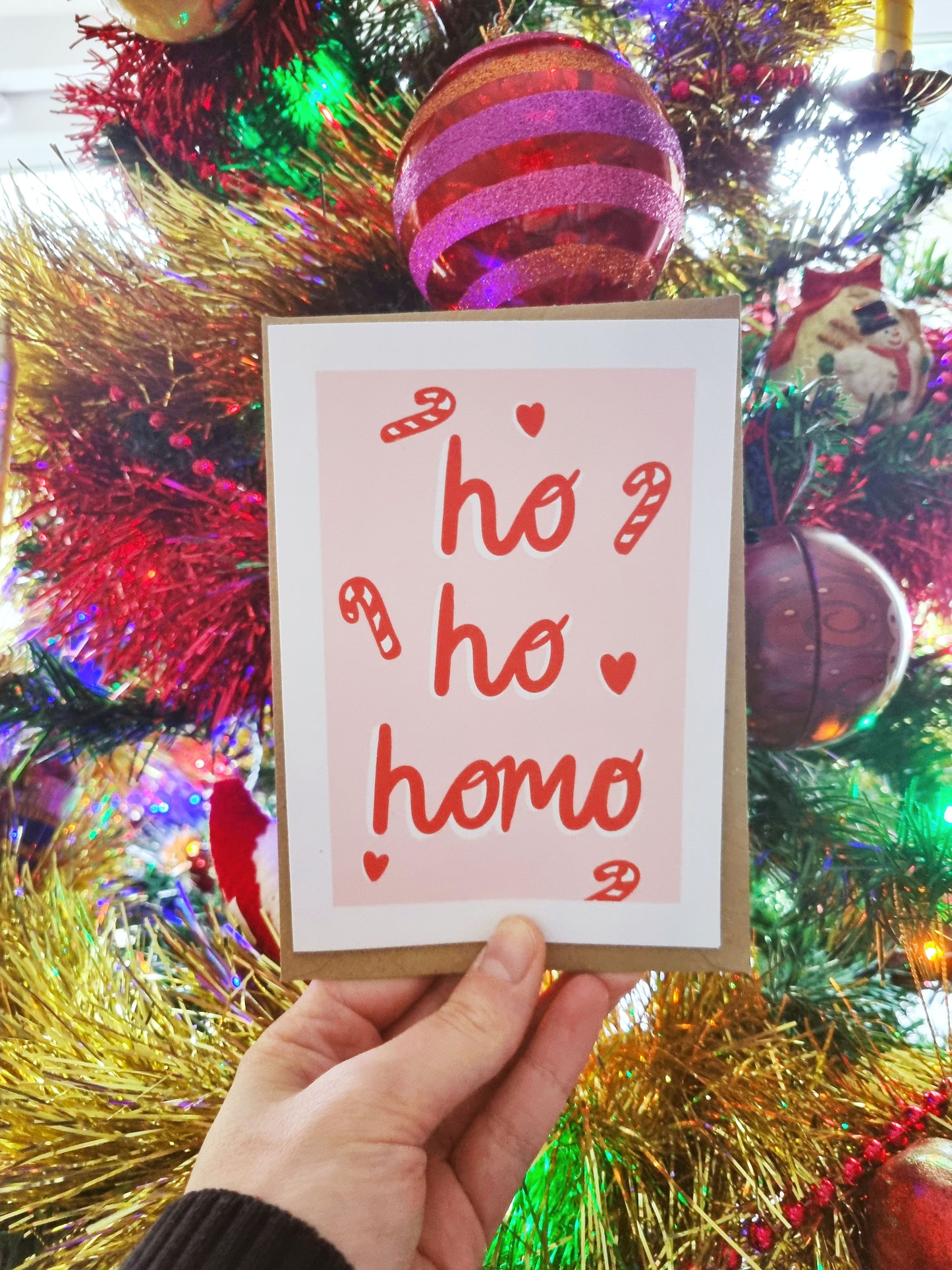 In front of a Christmas tree is a hand holding an A6 greeting card and kraft envelope. The card has a pastel pink rectangle with red candy canes and the phrase: Ho, Ho, Homo written in a cursive font.
