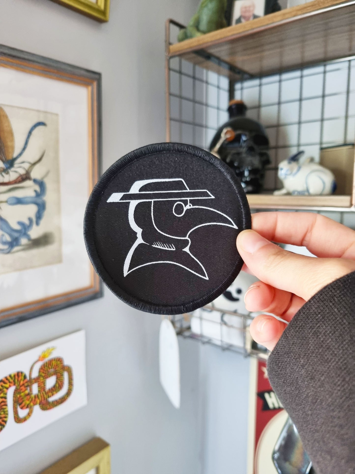 A hand holds a circler, black iron on patch with an overlocked border on the outside of the circle. Inside the patch in white ink is a simple plague doctor motif.