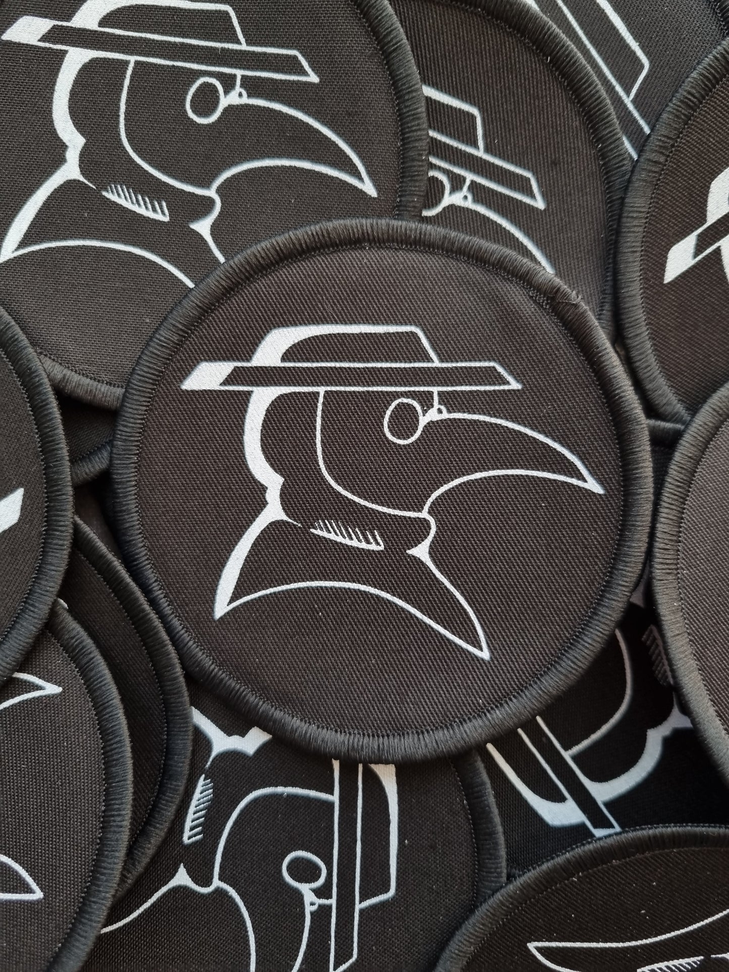 In the centre of the photo is a black, circular, cotton patch. The patch has an overlocked border. Inside the circle is a simple plague doctor design, printed in white ink. Underneath this patch is multiple of the same patch filling the screen.