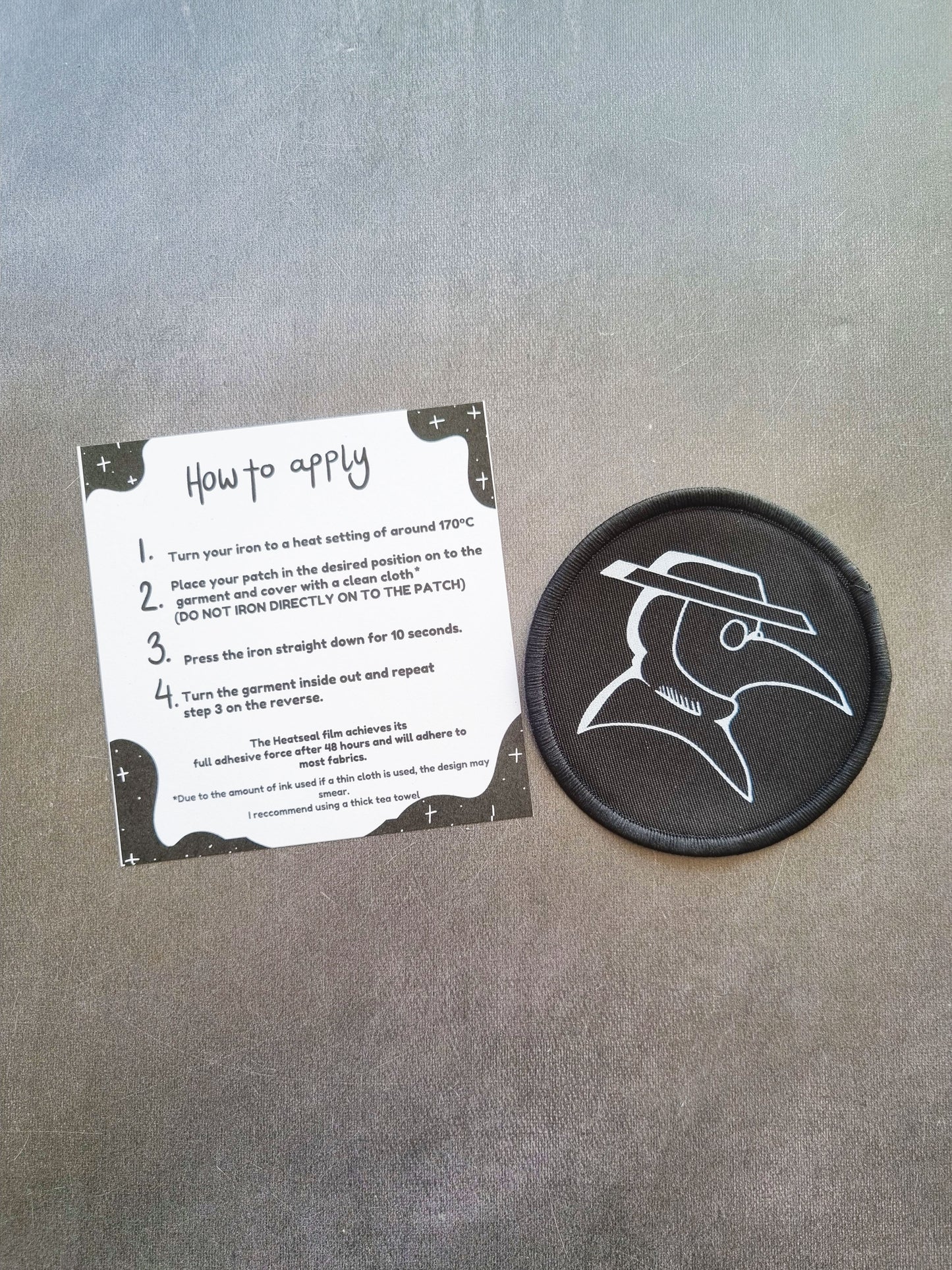 On a grey background, an instruction sheet of how to apply the iron on patch lays next to a circler iron on patch with a plague doctor design.