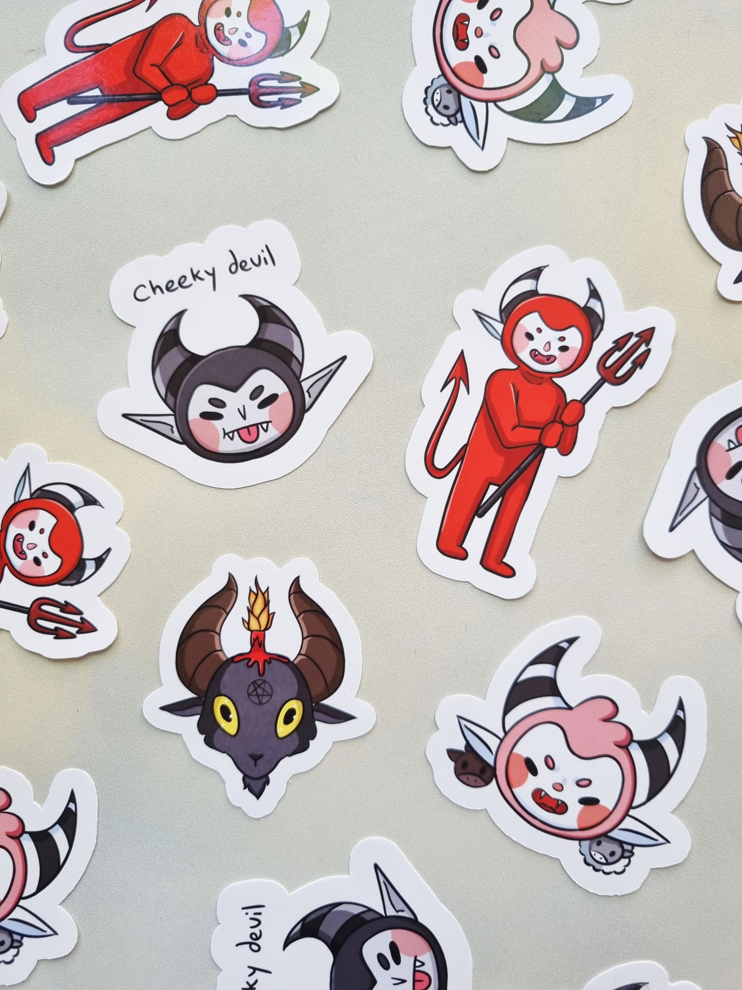 On a grey background stickers are scattered. They consist of: A cute-chibi style red devil holding a pitchfork, Baphomet, a pink devil with a cow and sheep, and a grey devil with the phrase "cheeky devil" above it.