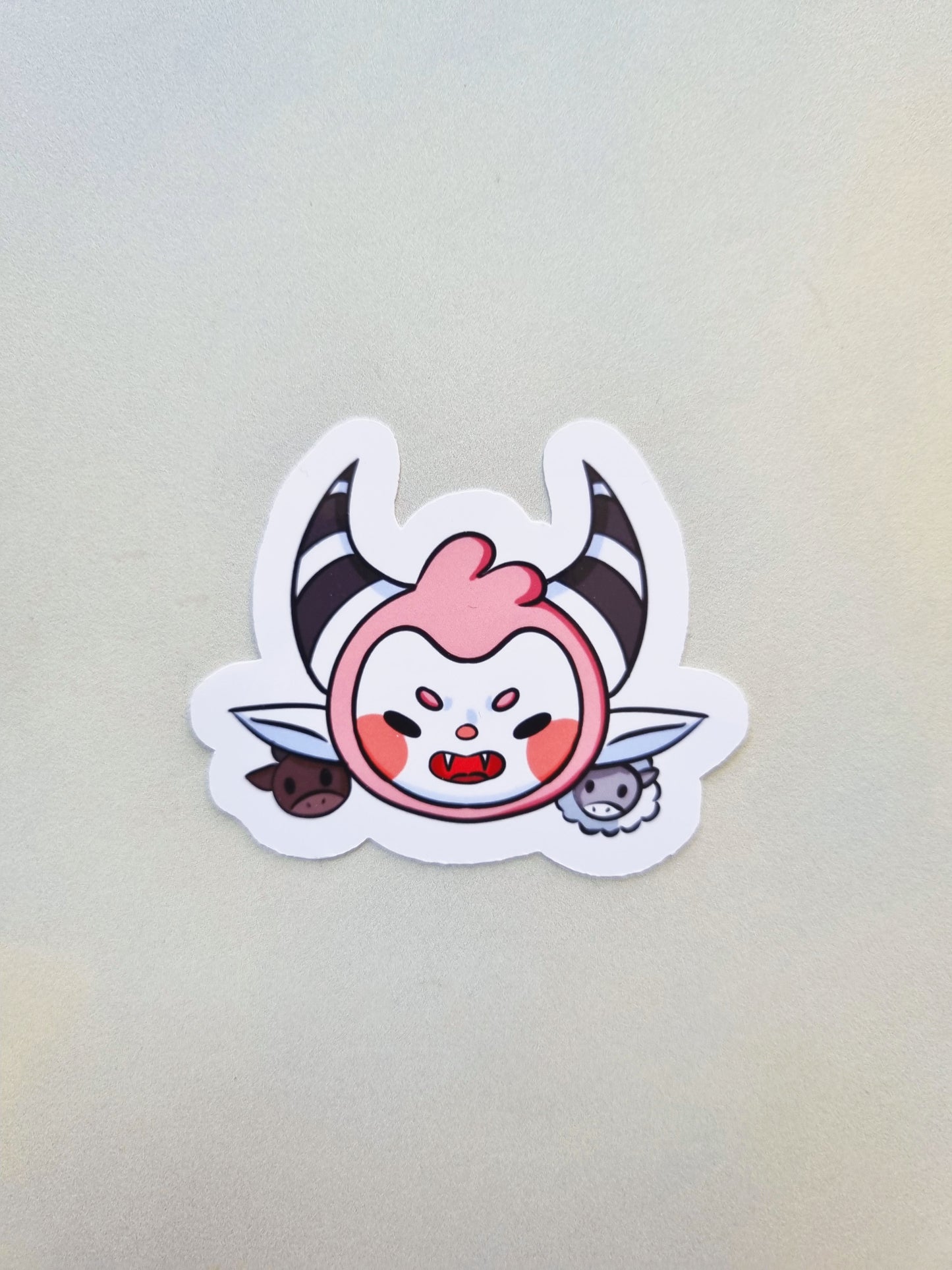 Sticker of a pink devil with black and white horns, with a cow and sheep besides it.