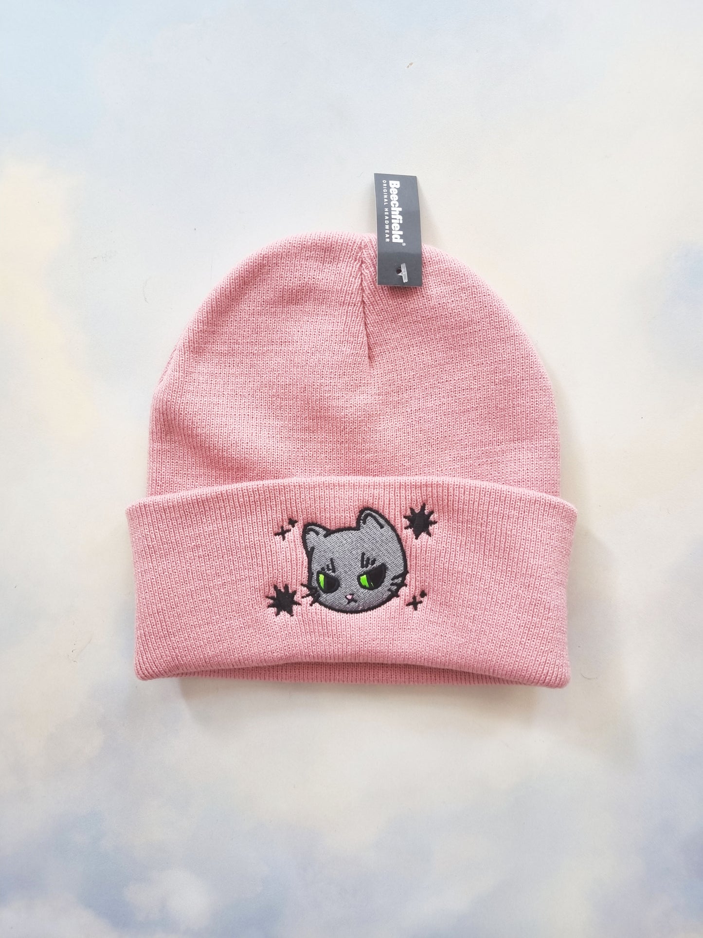 A pink beanie on a sky background, with a grey cat design embroidered onto it. The cat has a grumpy expression. Around the cat design is also black stars.