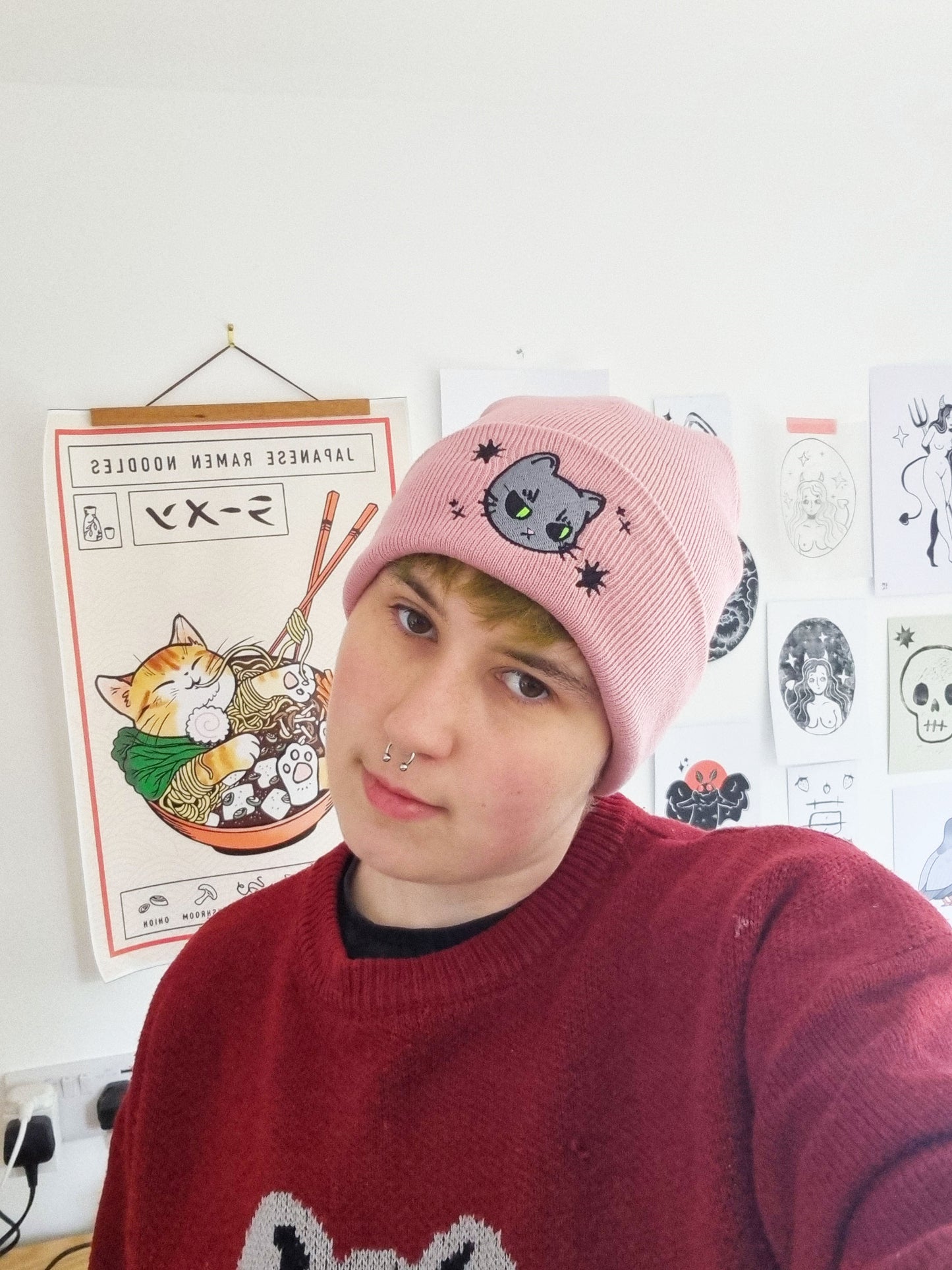 A person wearing a pink beanie with a grey cat design embroidered onto it.