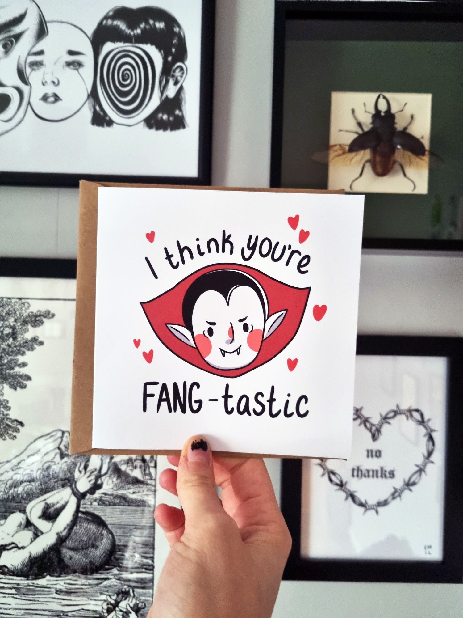 A hand holds a square greeting card that reads: I think you're fang-tastic. The wording is around a cute vampire with a red cape collar and red hearts are surrounding the vampire.