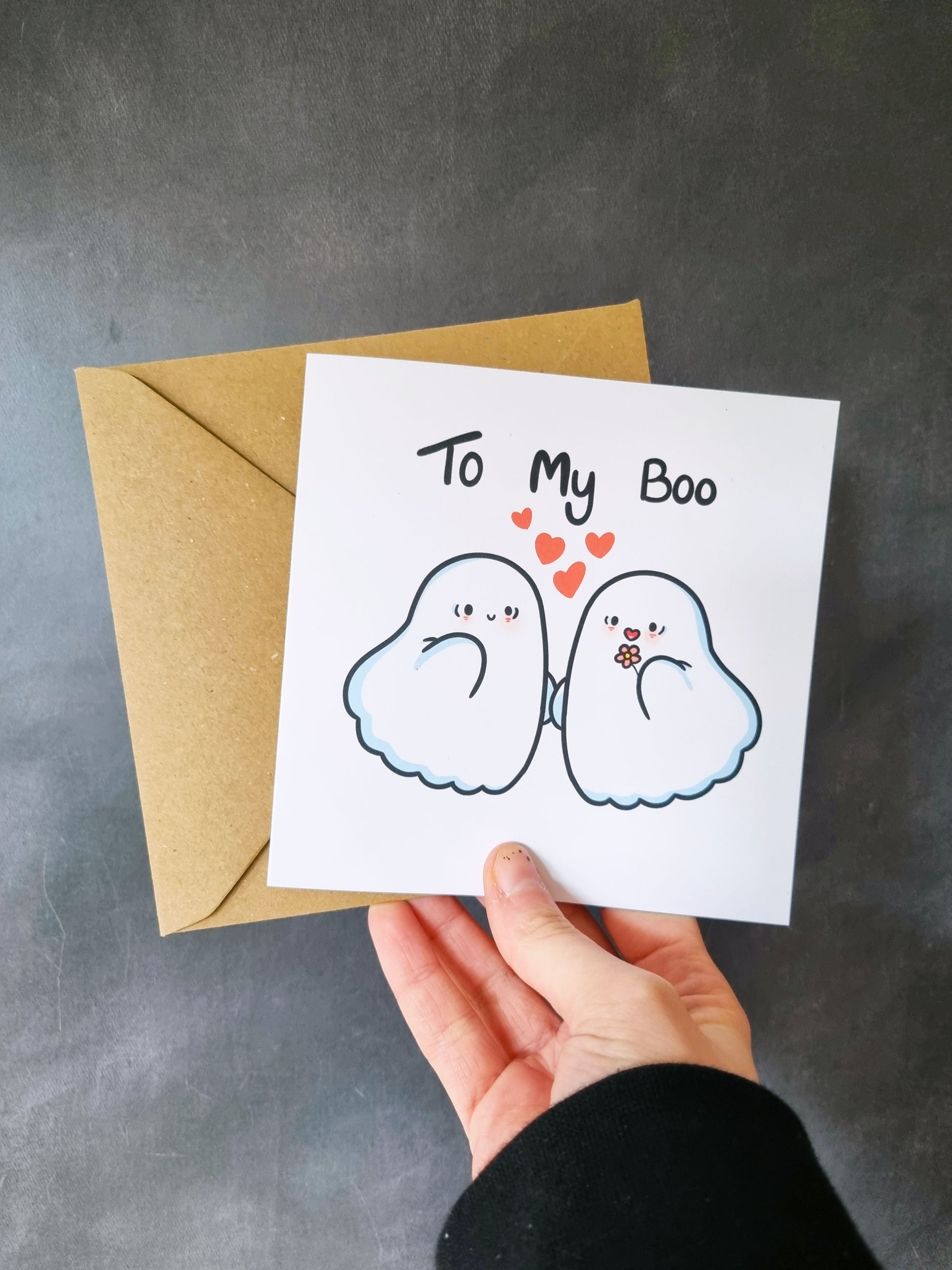 A hand holds a greeting card in front of a grey background. On the square greeting card has two cute ghosts, one holding a flower with 4 hearts in between the two ghosts. Above them reads: To my boo. Behind the card is a kraft envelope.