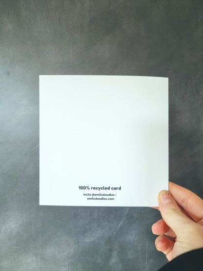 The back of a square greeting card being held in front of a grey background. The only writing is the branding of the website.