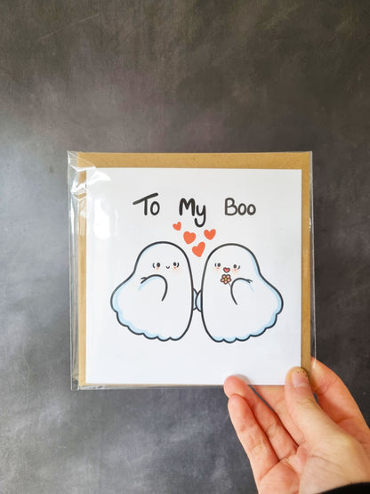A packaged greeting card in a clear bag being held in front of a grey background. The card has two cute ghosts with the phrase: To my boo, above them.