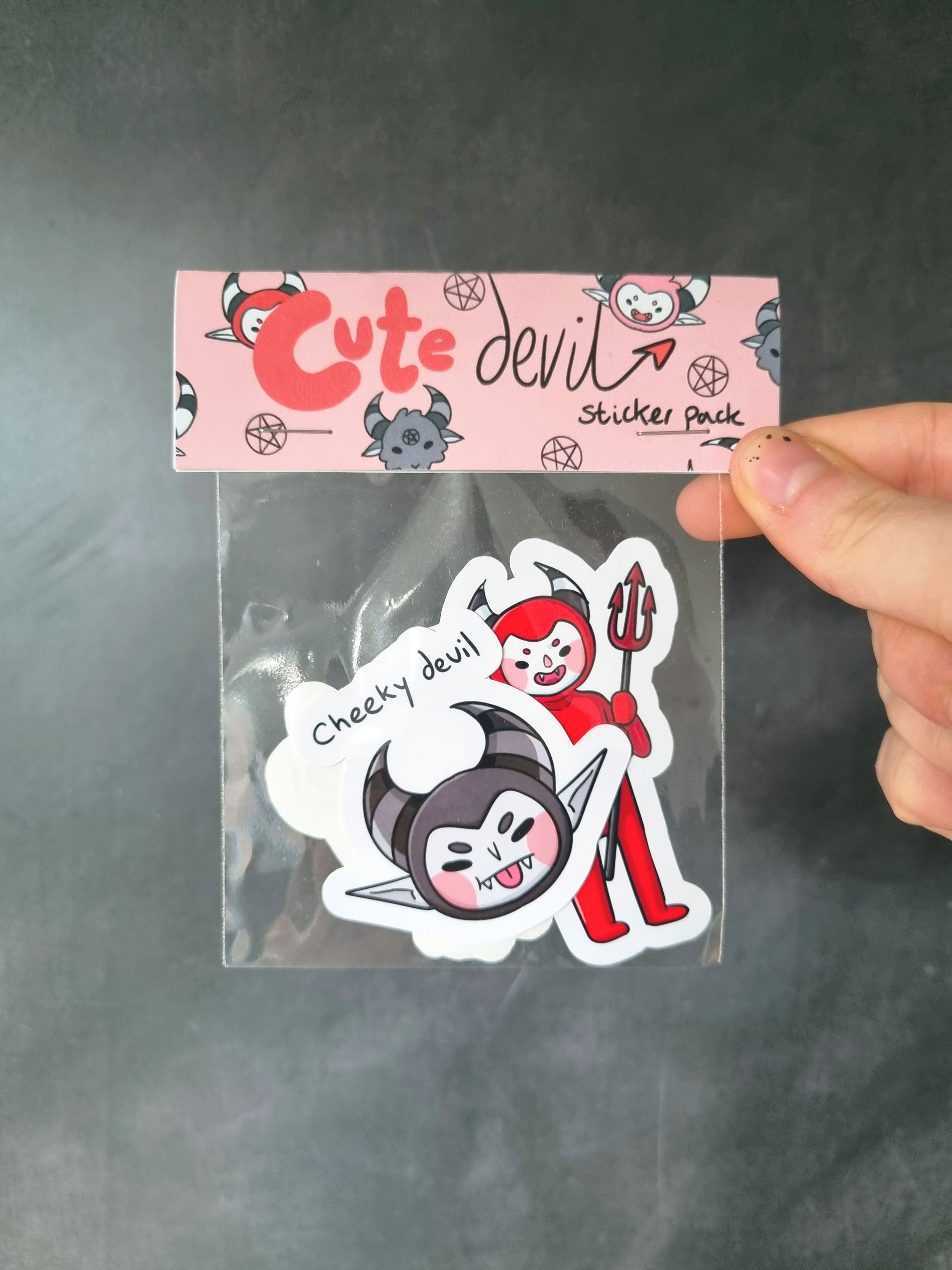 In front of a grey background a hand holds a sticker pack. The header of the sticker pack is baby pink with repeating cute devils and pentagrams. The header in different fonts reads, Cute Devil sticker pack. In a clear bag is several stickers.