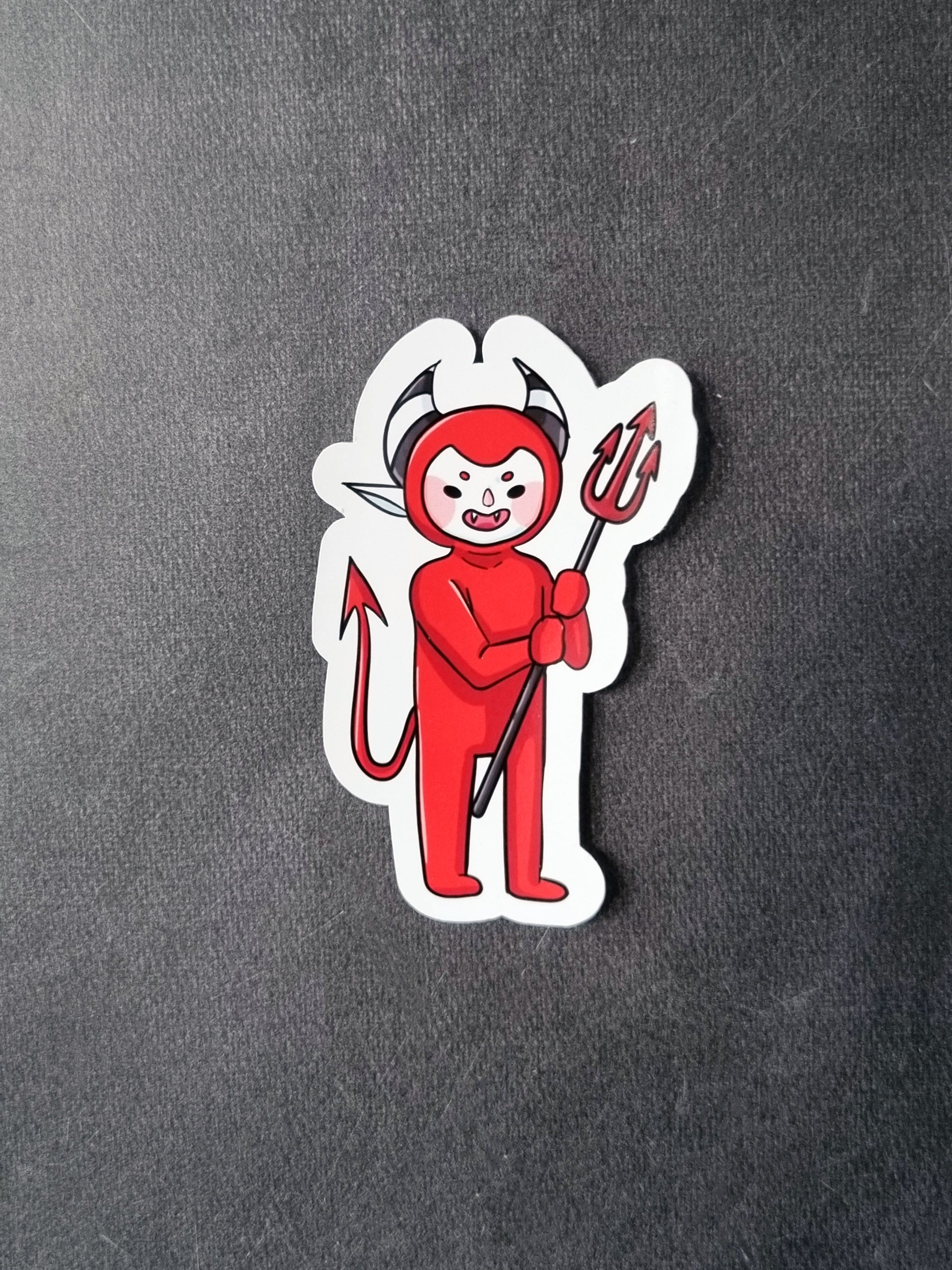 On a grey background is a glossy sticker. The sticker is a devil in a red full body suit leaving the face open. The devil is holding a pitchfork. He has black and white horns and baby pink blushed cheeks.