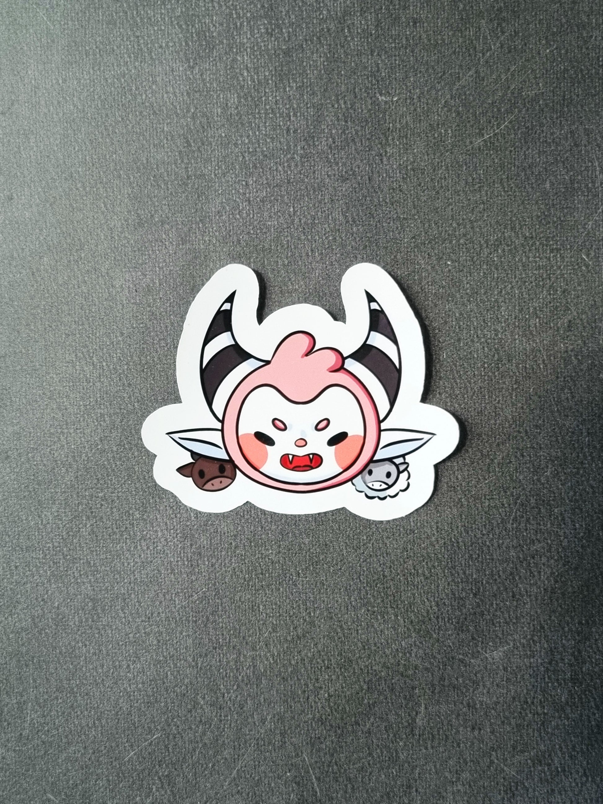 On a grey background, a glossy sticker is laid. The sticker is the face of a cute devil with pastel pink hair around a white face with pink blush. It has black and white horns, between those is a tuft of hair. Behind the pointed ears is a cow and sheep.