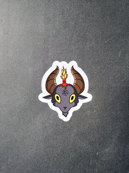 On a grey background, a cute sticker of a goat head with a pentagram on its forehead, and a red candle on top of his head between brown horns.