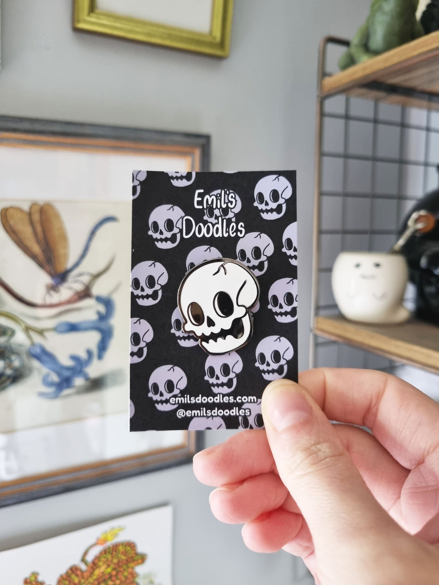 A hand holds a backing card with a skull enamel pin pinned to it. The skull pin is a cartoony design with a crack on the top of the skull. The backing card has the same design in an alternating pattern, with the shop name and social media handles.