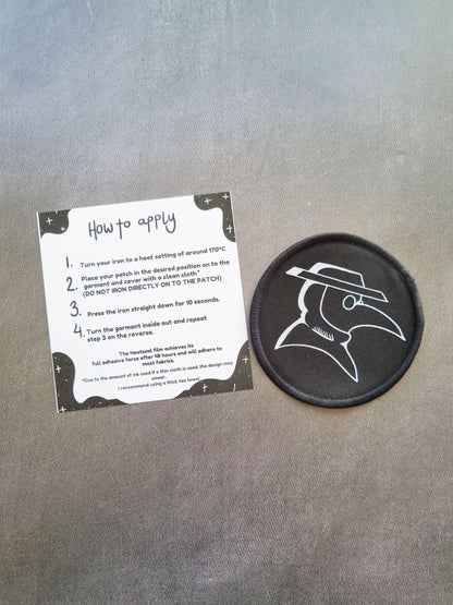 On a grey background, an instruction sheet of how to apply the iron on patch lays next to a circler iron on patch with a plague doctor design.
