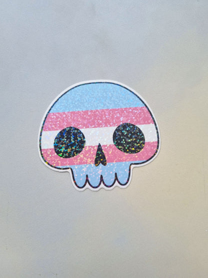 On a light background is a skull shaped sticker with the transgender pride flag inside. With a holographic overlay.