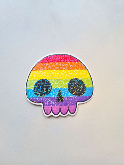 On a light background, is a sticker in the shape of a skull with the rainbow pride flag inside with a holographic overlay.