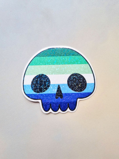 On a light background is a cartoony-skull shaped sticker with the gay pride flag inside the design. It has a holographic overlay.