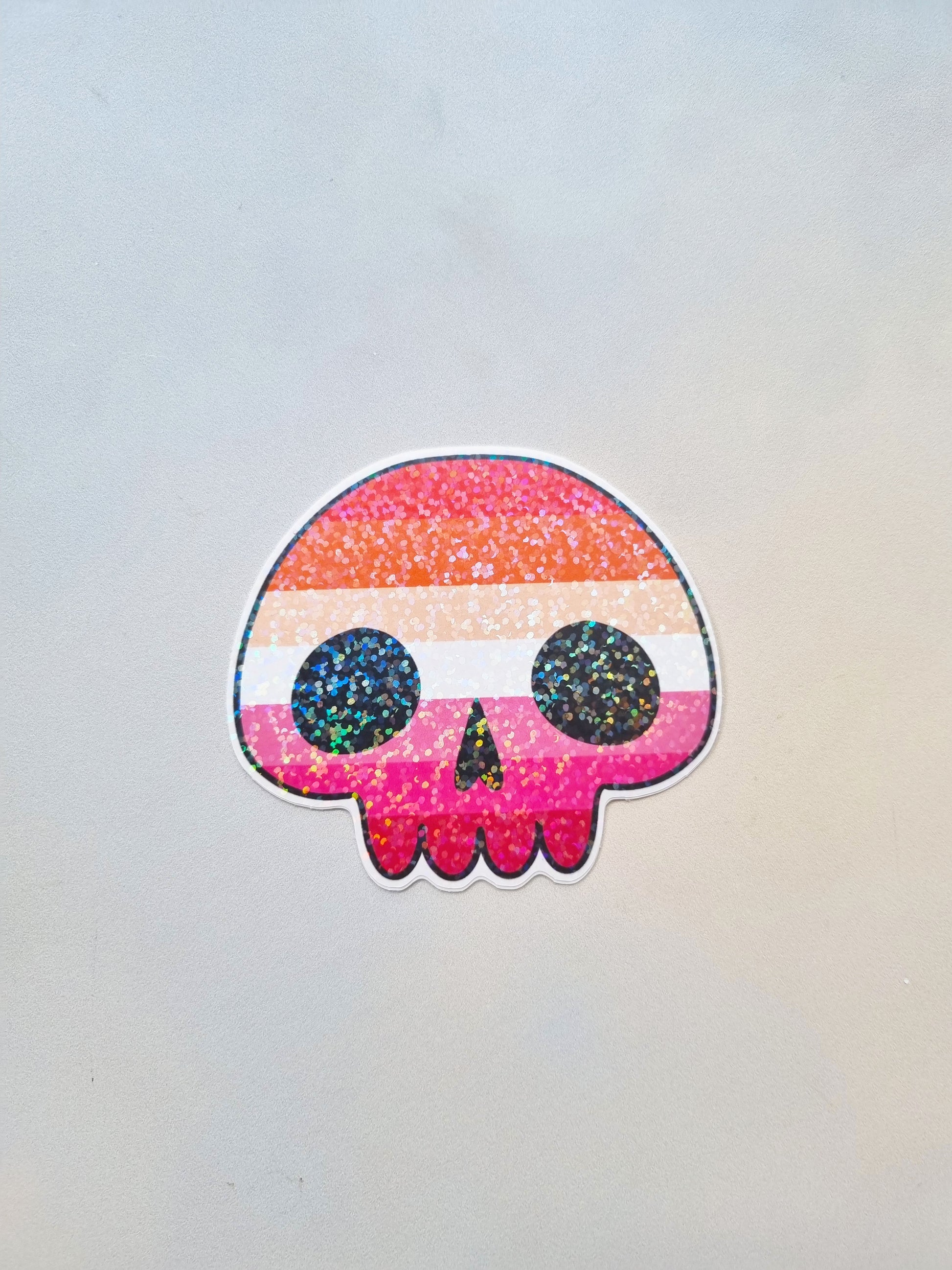 On a light background is a skull shaped sticker with the lesbian pride flag inside the flag. It has a holographic overlay.