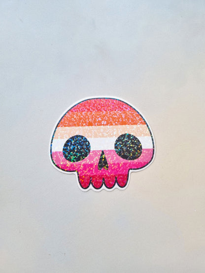 On a light background is a skull shaped sticker with the lesbian pride flag inside the flag. It has a holographic overlay.
