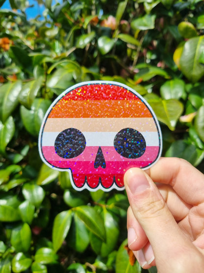 Outside in front of leaves, a hand is holding a lesbian pride flag sticker in the shape of a skull. It has a holographic  layer over it.