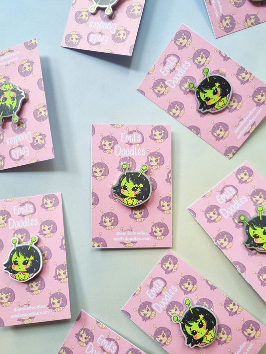 A cute alien character in a chibi style.  The alien is light green with black hair and eyes, wearing a star choker style necklace and antennas on top of her head. The alien pin is pinned on to a pink backing card with a repeating pattern of the same alien design.