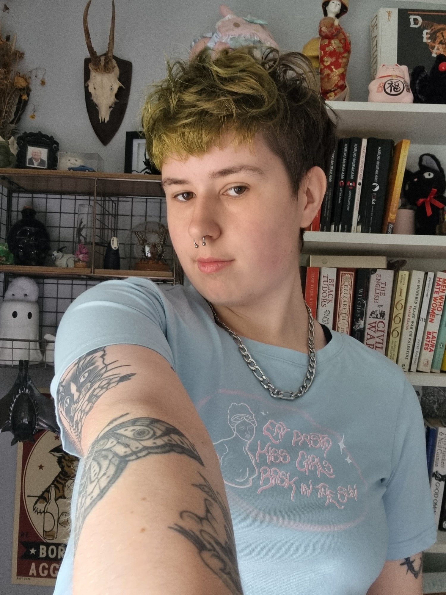Person with tattoos wearing a light blue t-shirt in a room with shelves and decor