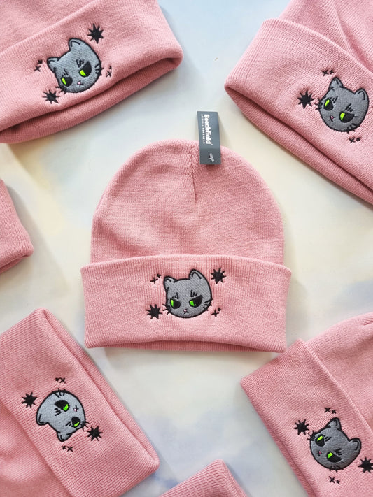 Pink beanies on a sky background. Each beanie has a grey, grumpy cat design embroidered on it. Around the cat design is also black stars.