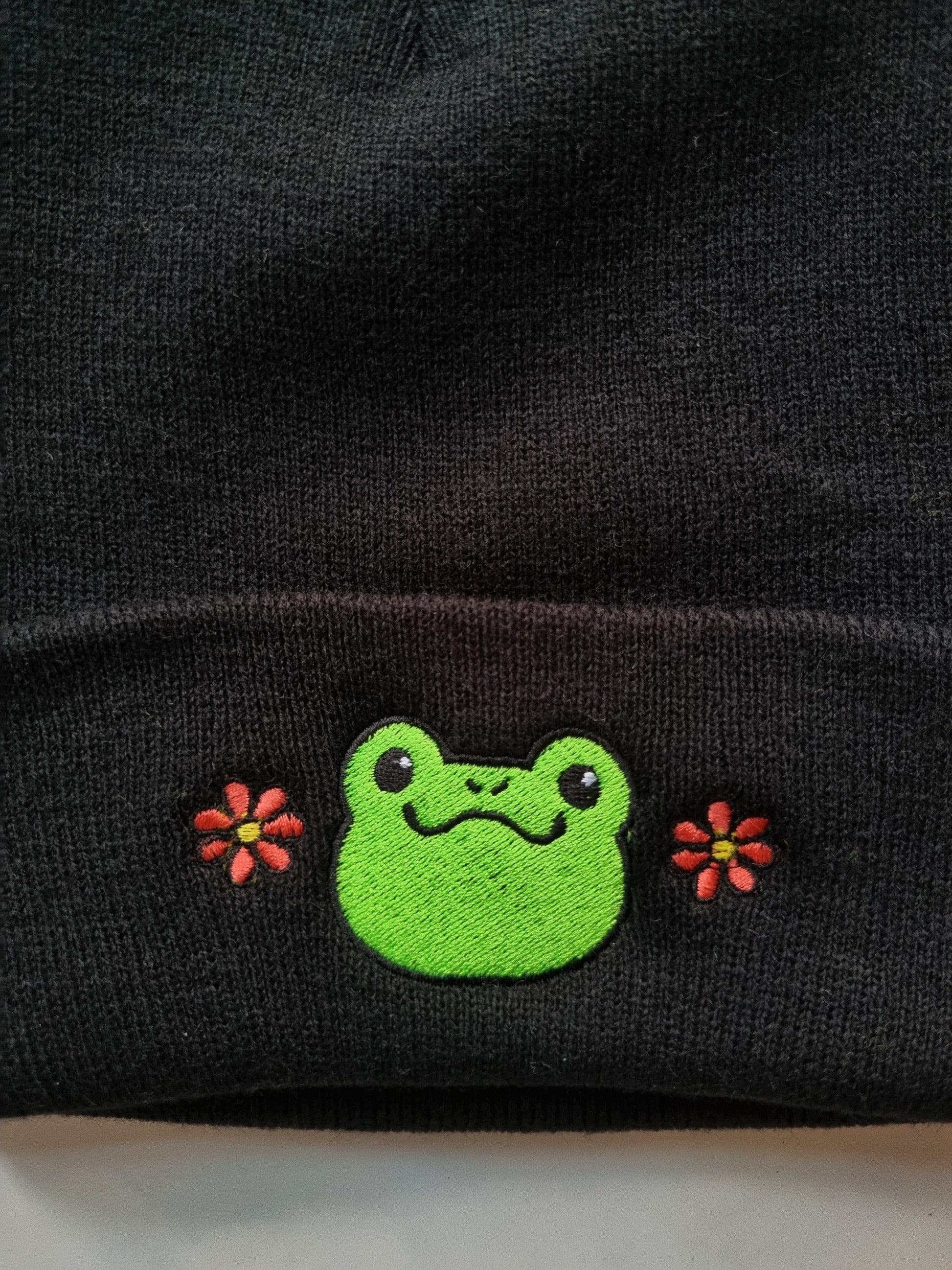 A black beanie with a embroidered design of a cute, green frog face that has two red flowers on each side.